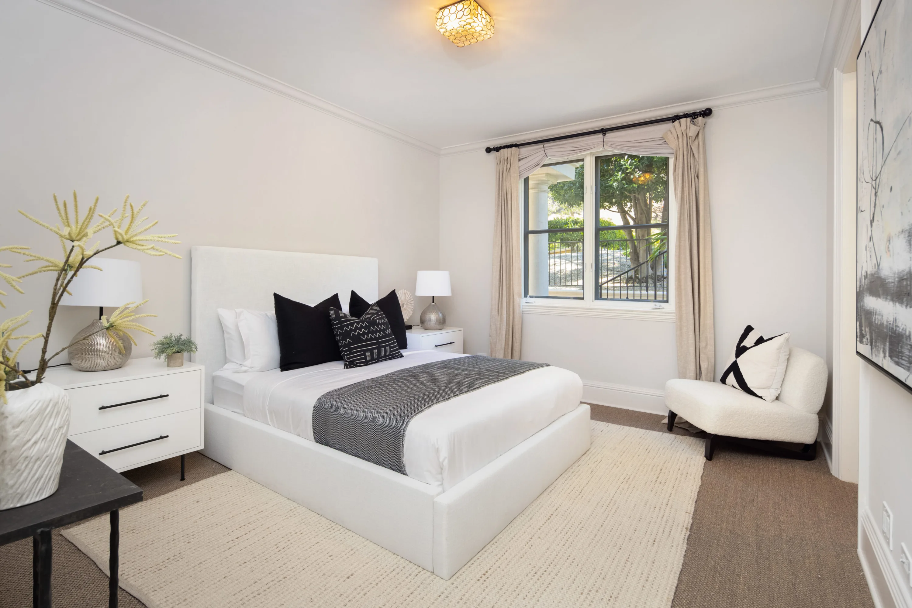 Bright bedroom with white headboard, black and cream cushions, bouclé chair and treetops beyond the window