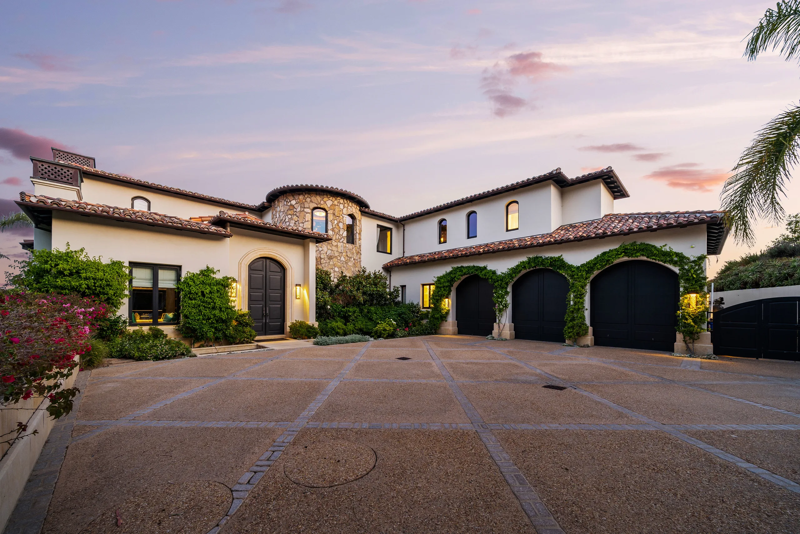 Arrival courtyard at twilight, arched front door and three ivy-draped garage bays under warm-lit stucco