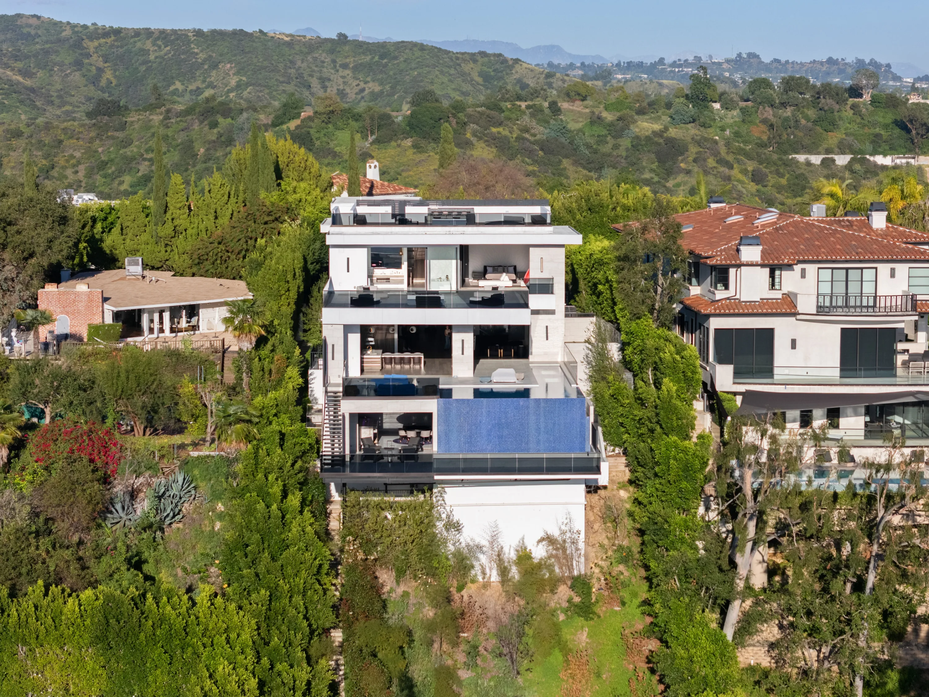 Aerial daytime view of the multi-level estate stacked above a deep blue glass-edged pool on the hillside
