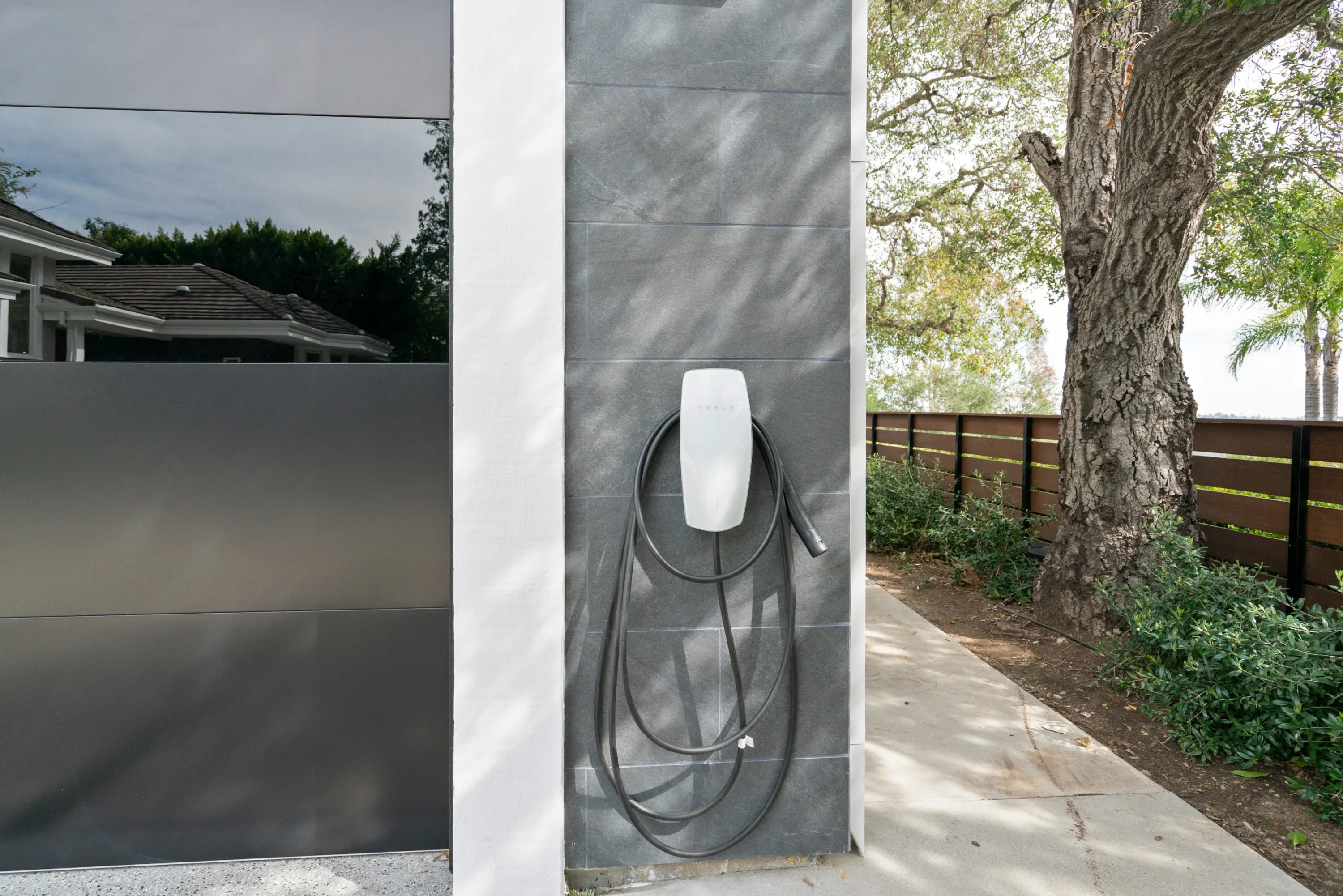 Wall-mounted electric car charger on stone cladding beside the garage door, with oak-shaded driveway alongside
