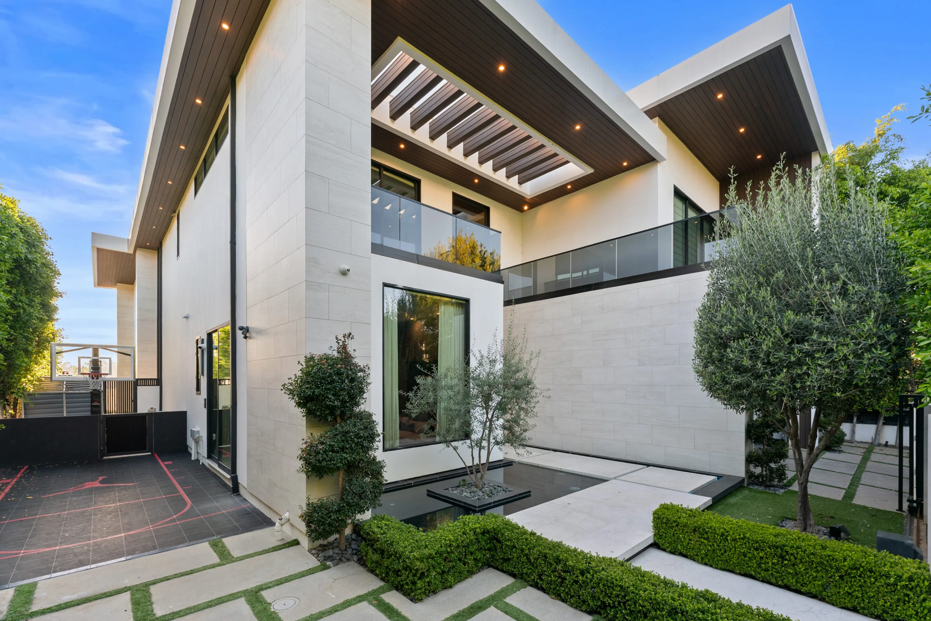 Stone-clad entry courtyard with reflecting pool, olive trees, clipped hedging and a painted sport court alongside