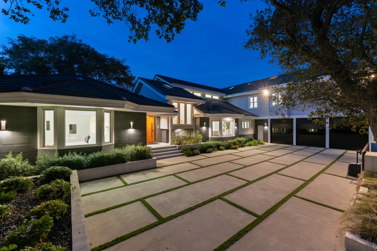 Front elevation at blue hour, warm-lit windows above a grass-jointed motor court leading to the timber entry door