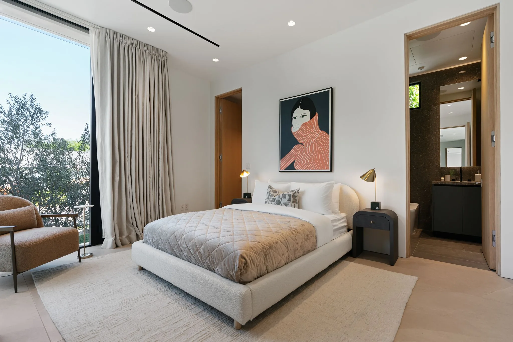 Bedroom with bouclé bed, brass reading lamps, portrait artwork and floor-to-ceiling glass onto olive trees