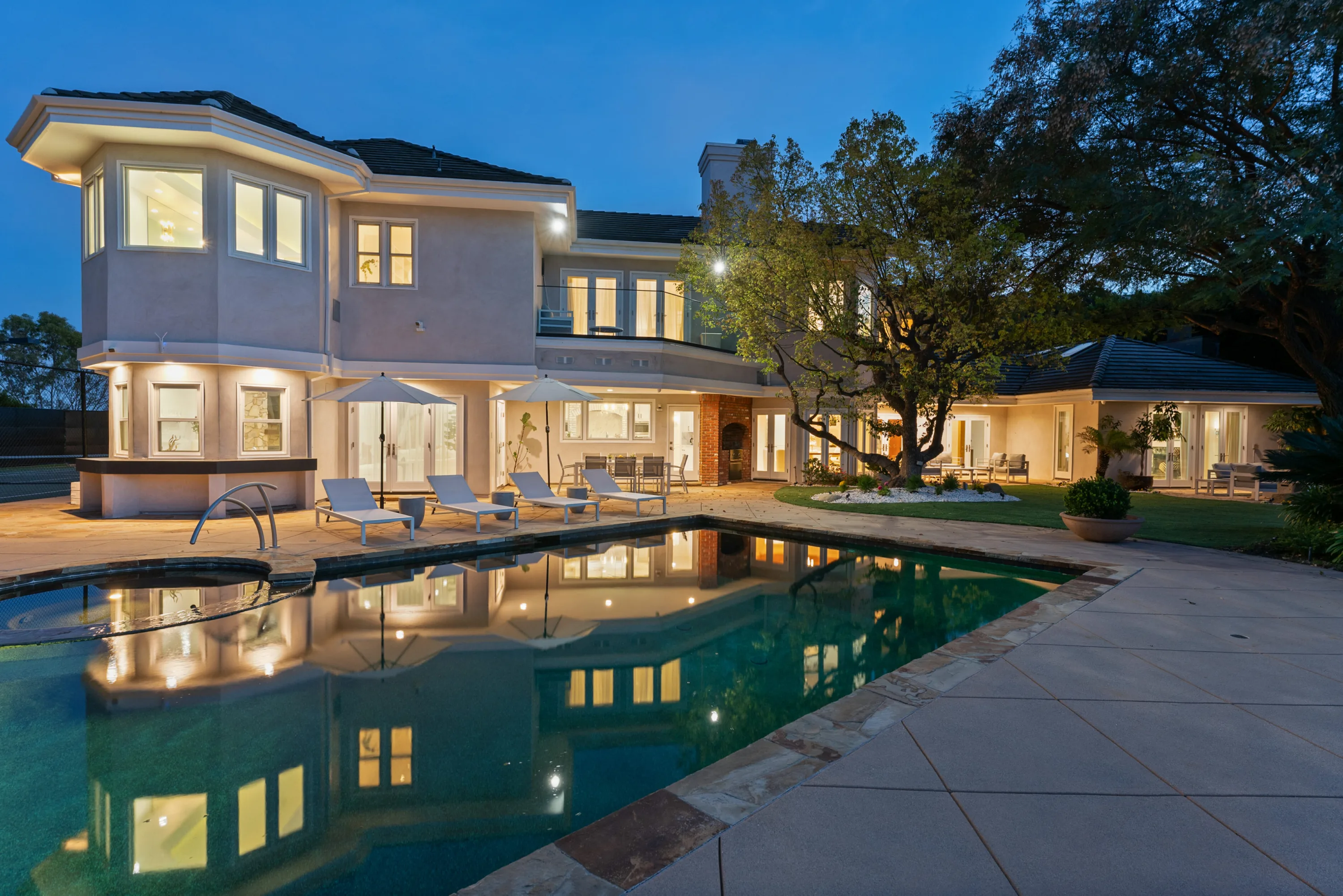 Rear facade mirrored in the pool at dusk, sun loungers, twin umbrellas and a spa set against the lit terrace