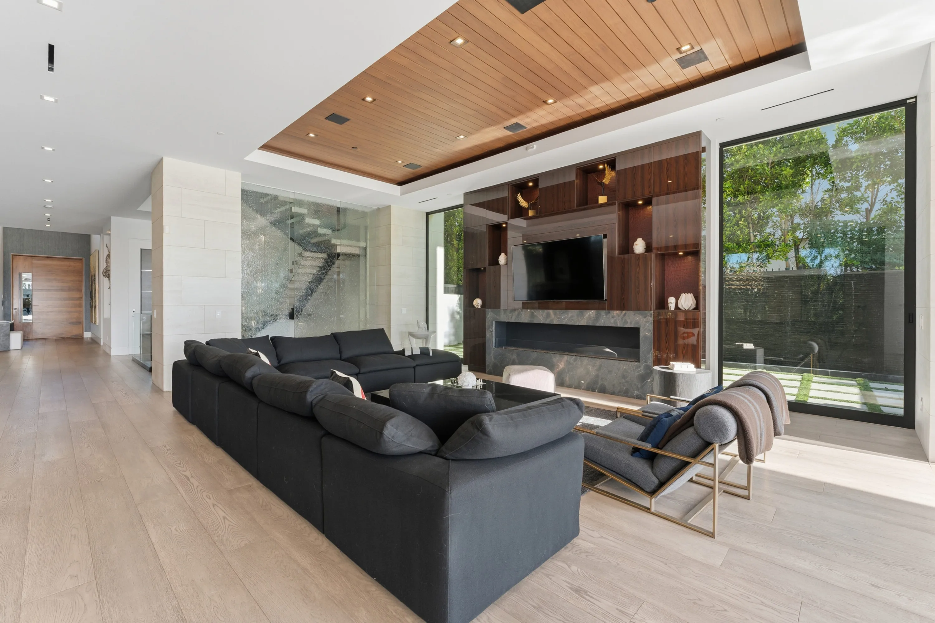 Second sitting room under a slatted wood ceiling, media wall above a stone fireplace and floating stair behind glass