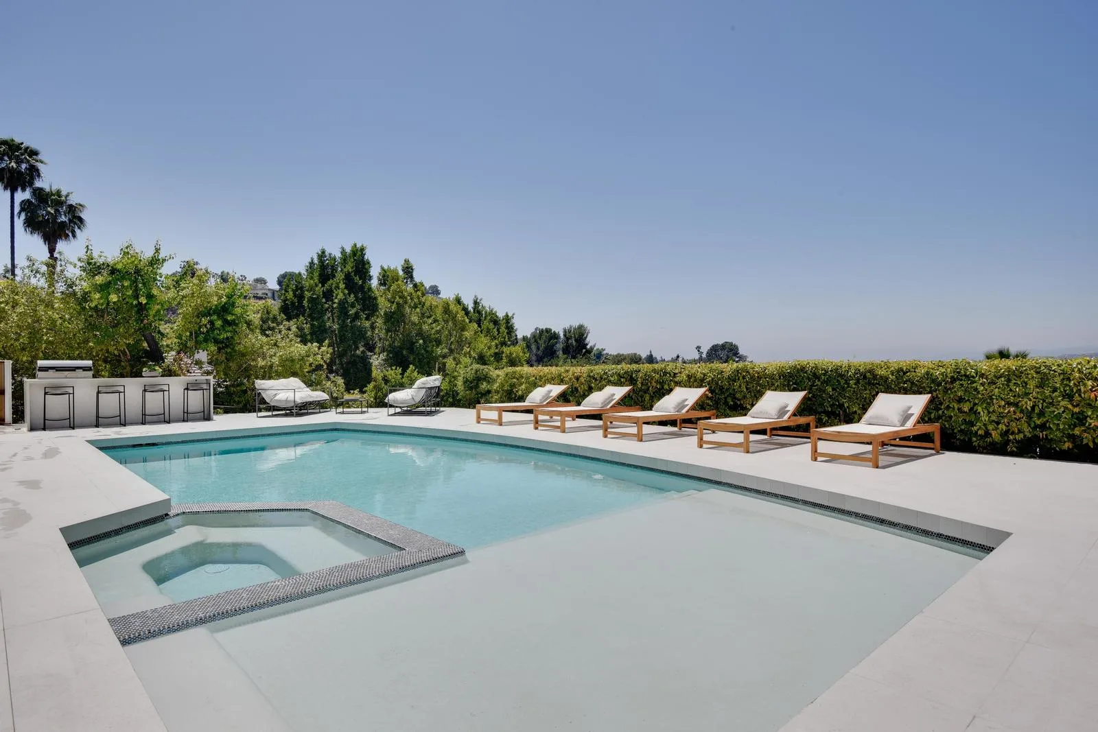 Private pool and inset spa edged by teak loungers, an outdoor bar and a clipped hedge under clear sky