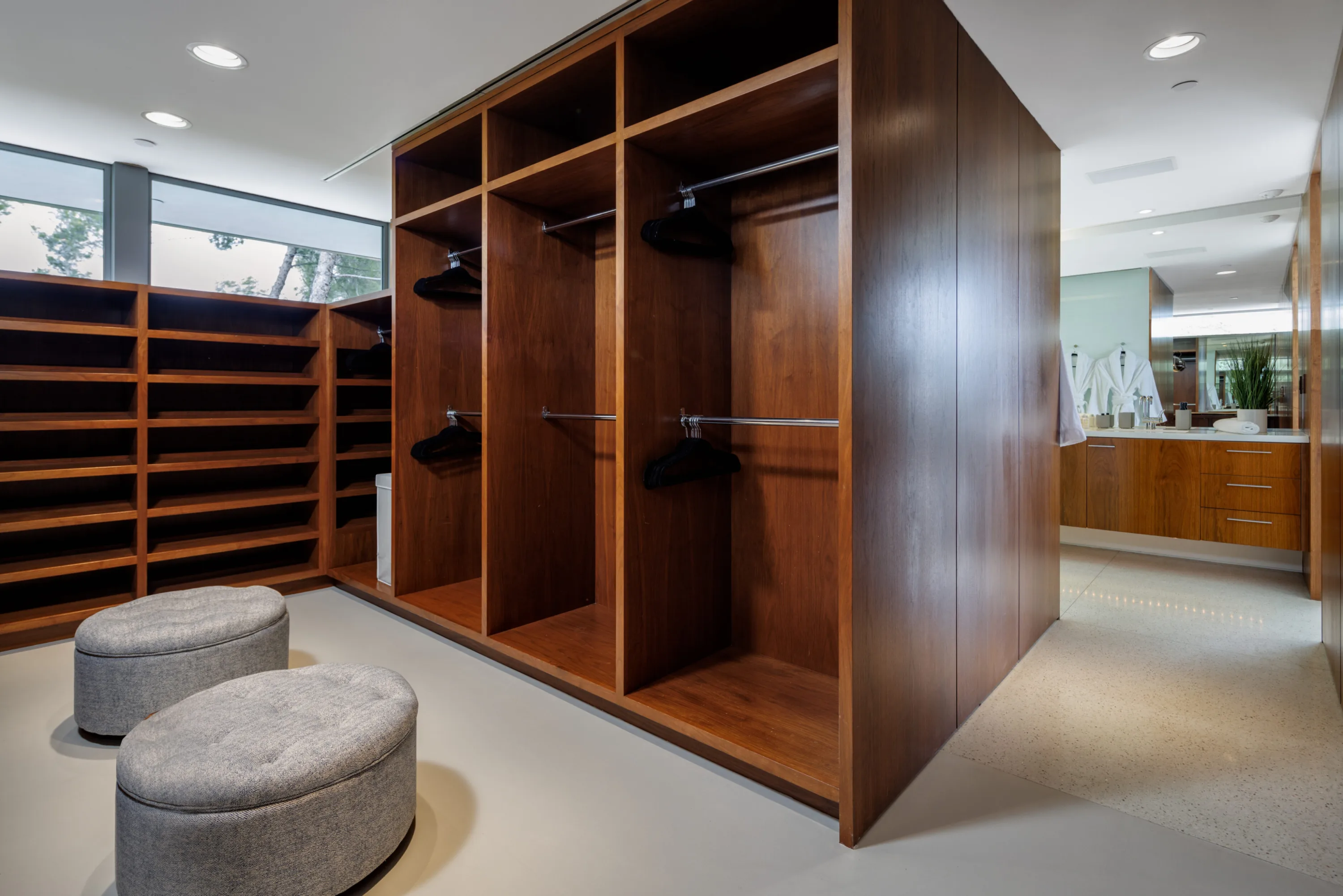 Walk-in dressing room in walnut with open hanging bays, shelving and grey ottomans, bath beyond