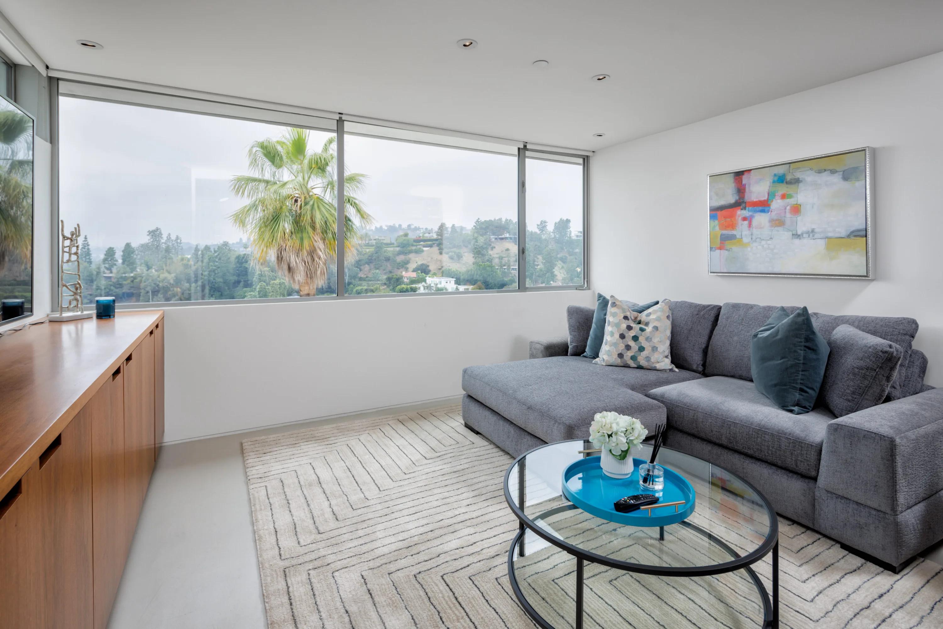 Sitting room with grey sectional, glass coffee table and corner windows framing palms and hillside homes