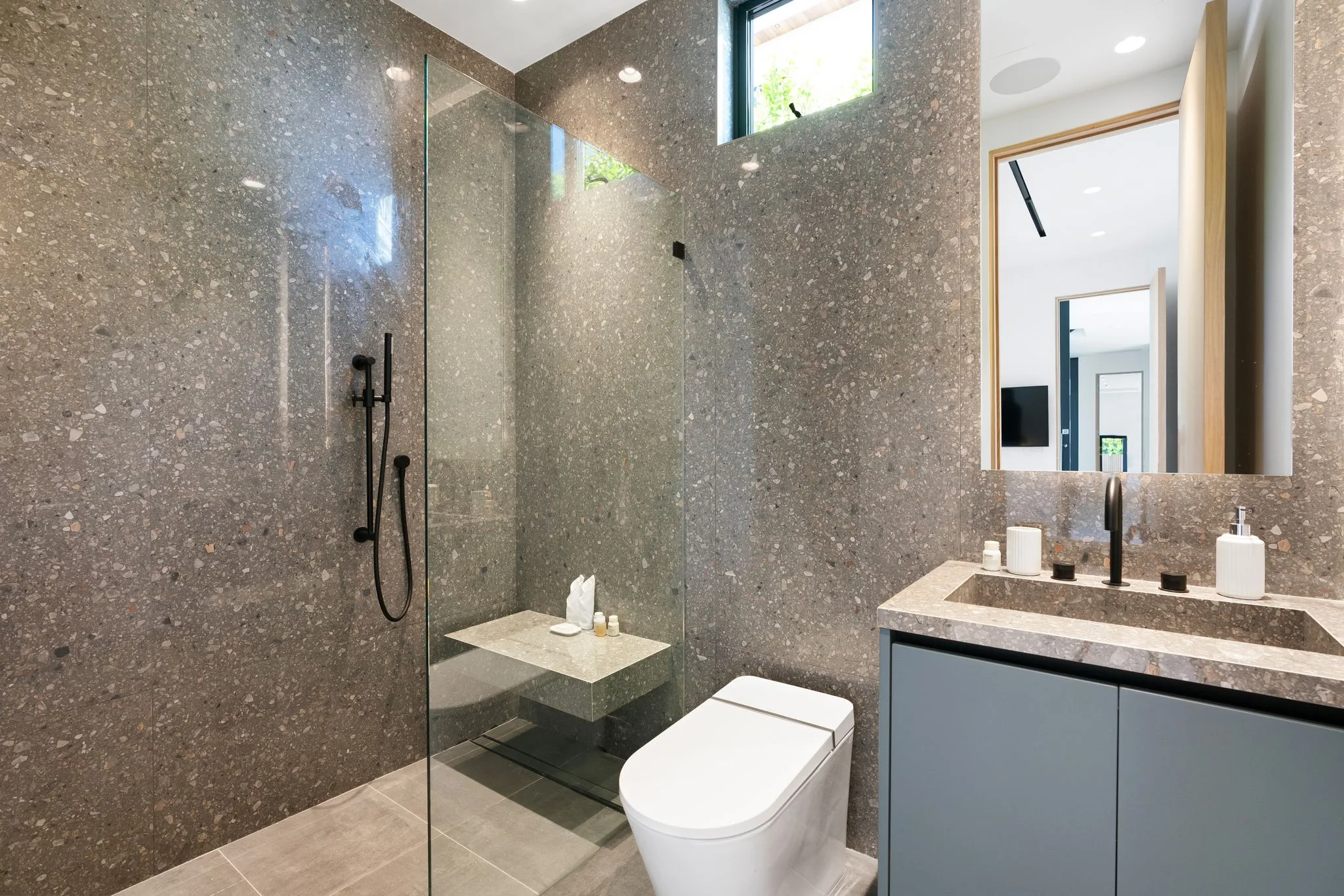 Terrazzo-clad guest bathroom with glass shower, stone bench and clerestory window framing greenery