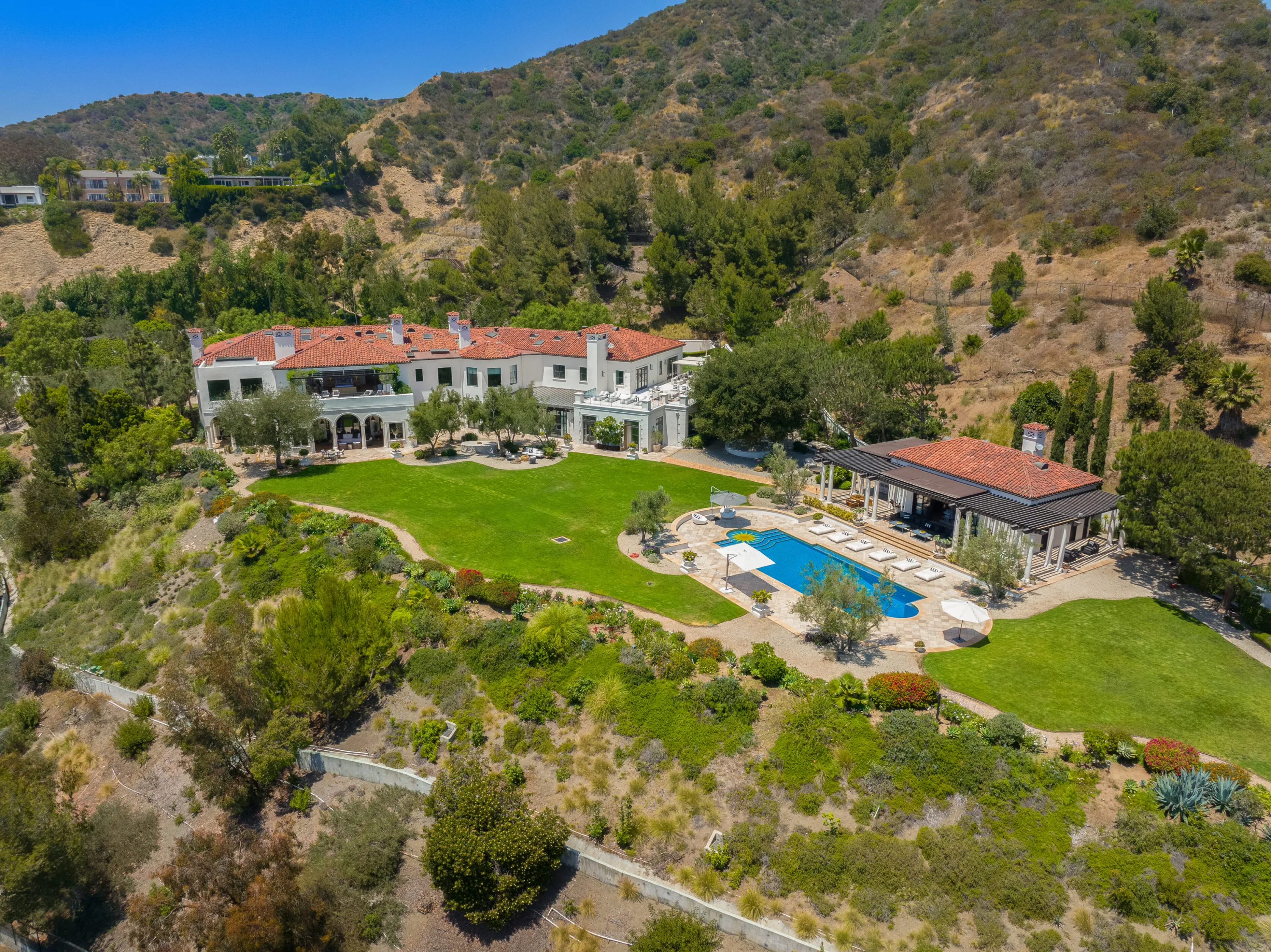 Aerial view of a gated Beverly Hills estate with red-tile roof, lawns, pool and poolside pavilion set into the hillside