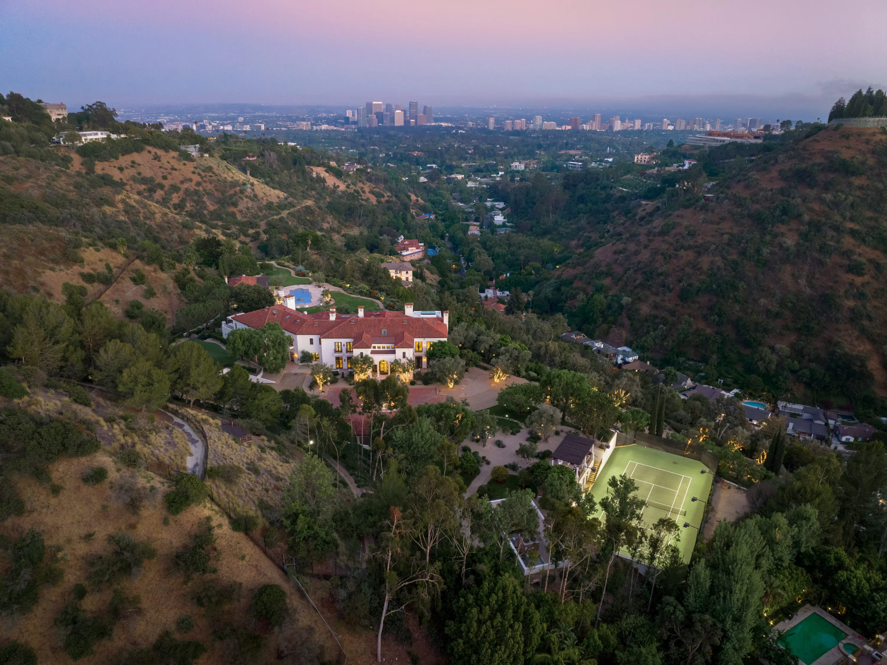 Dusk aerial of the lit estate and tennis court in the canyon, with the Century City skyline beyond