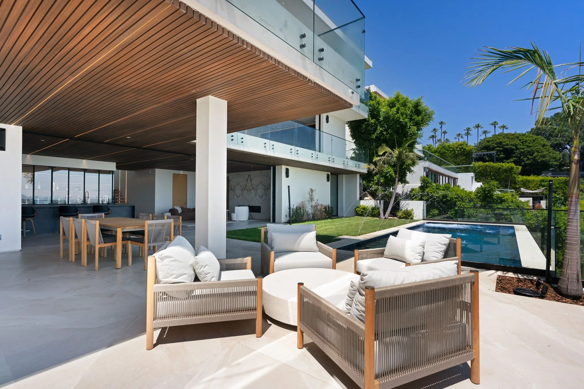 Shaded loggia seating cluster around a round stone table, lawn and pool stretching toward palm-lined hills