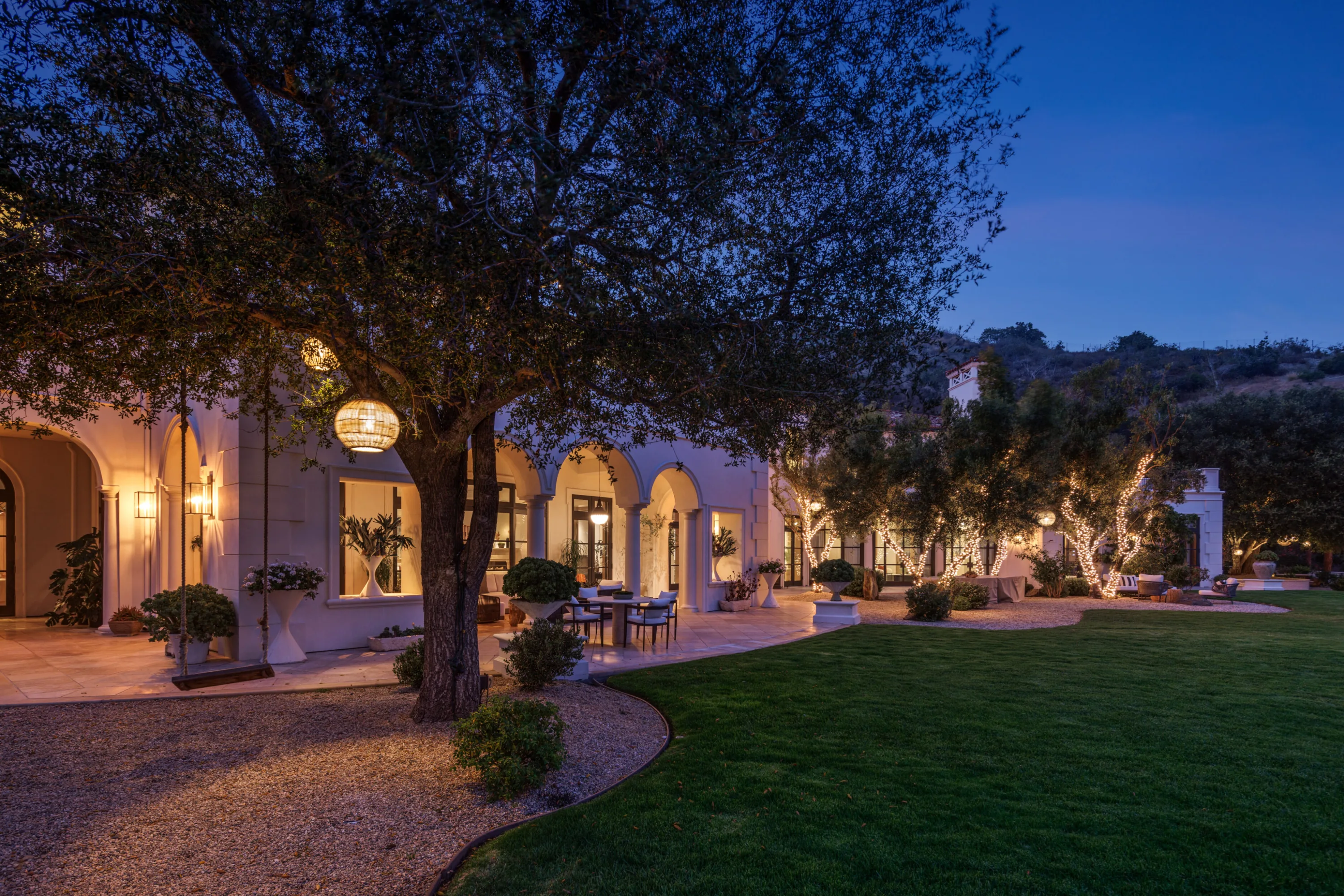 Arcaded rear elevation glowing at twilight beside the lawn, olive trees wrapped in fairy lights