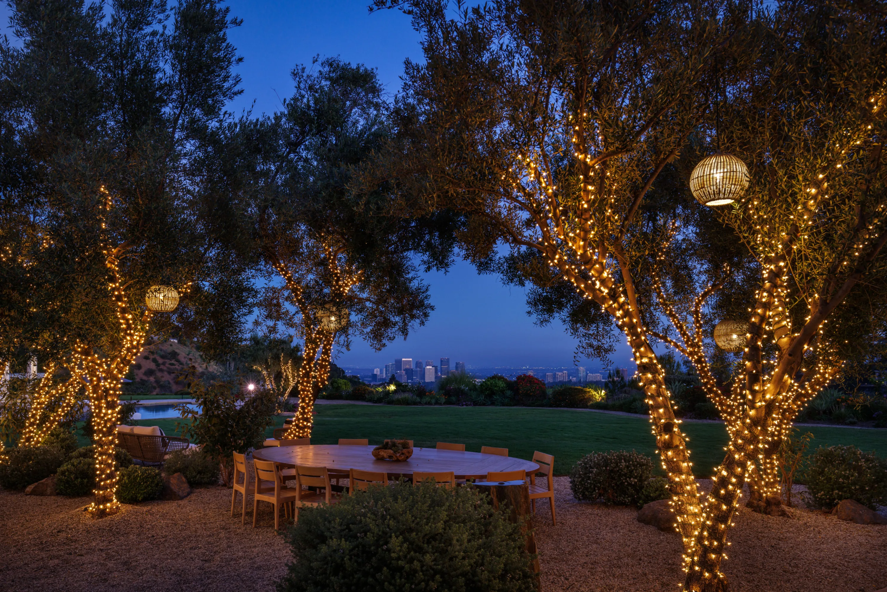 Round outdoor dining table under lantern-hung olive trees, city lights twinkling past the lawn