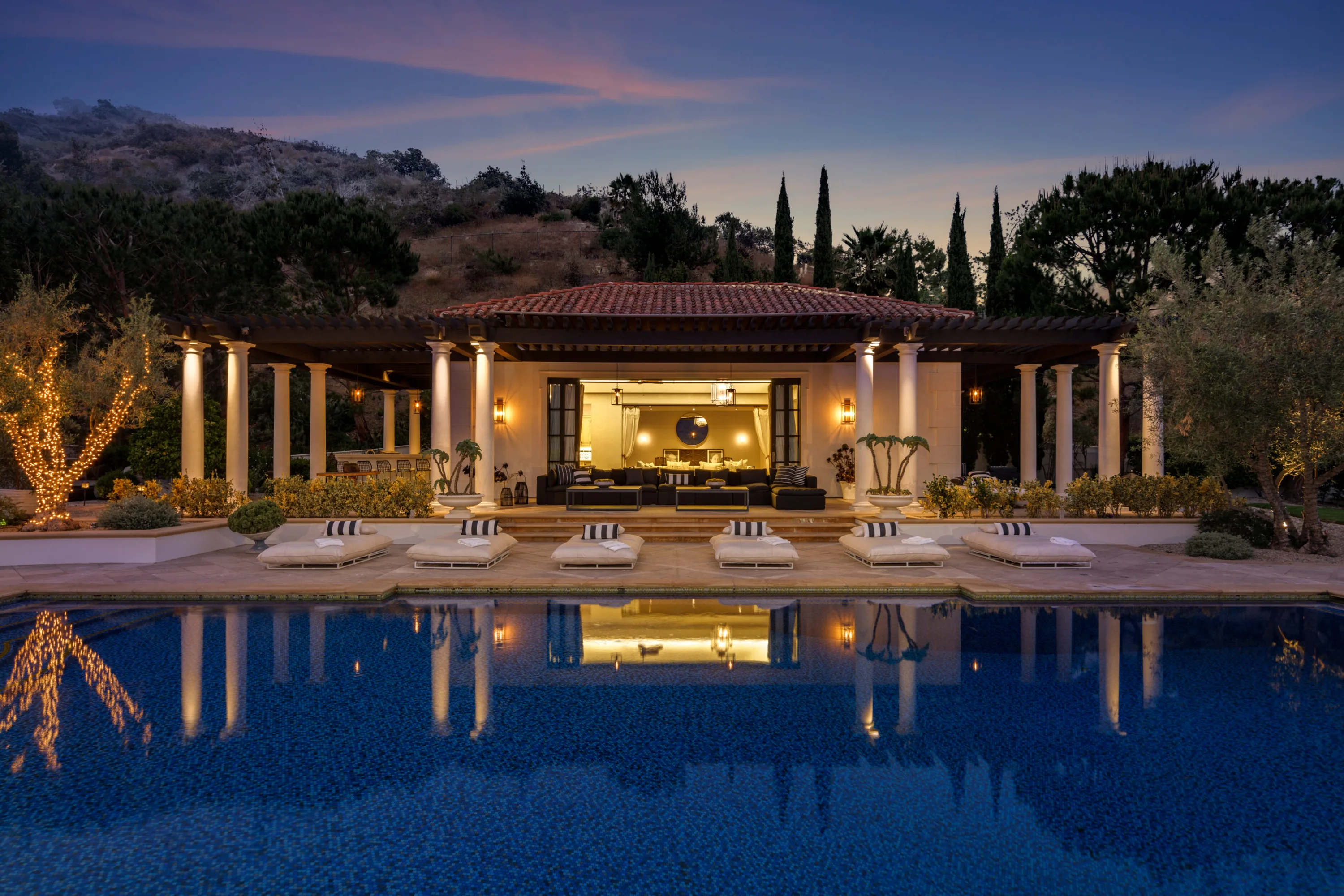 Colonnaded pool house lit at dusk, mirrored in the still swimming pool with striped loungers alongside