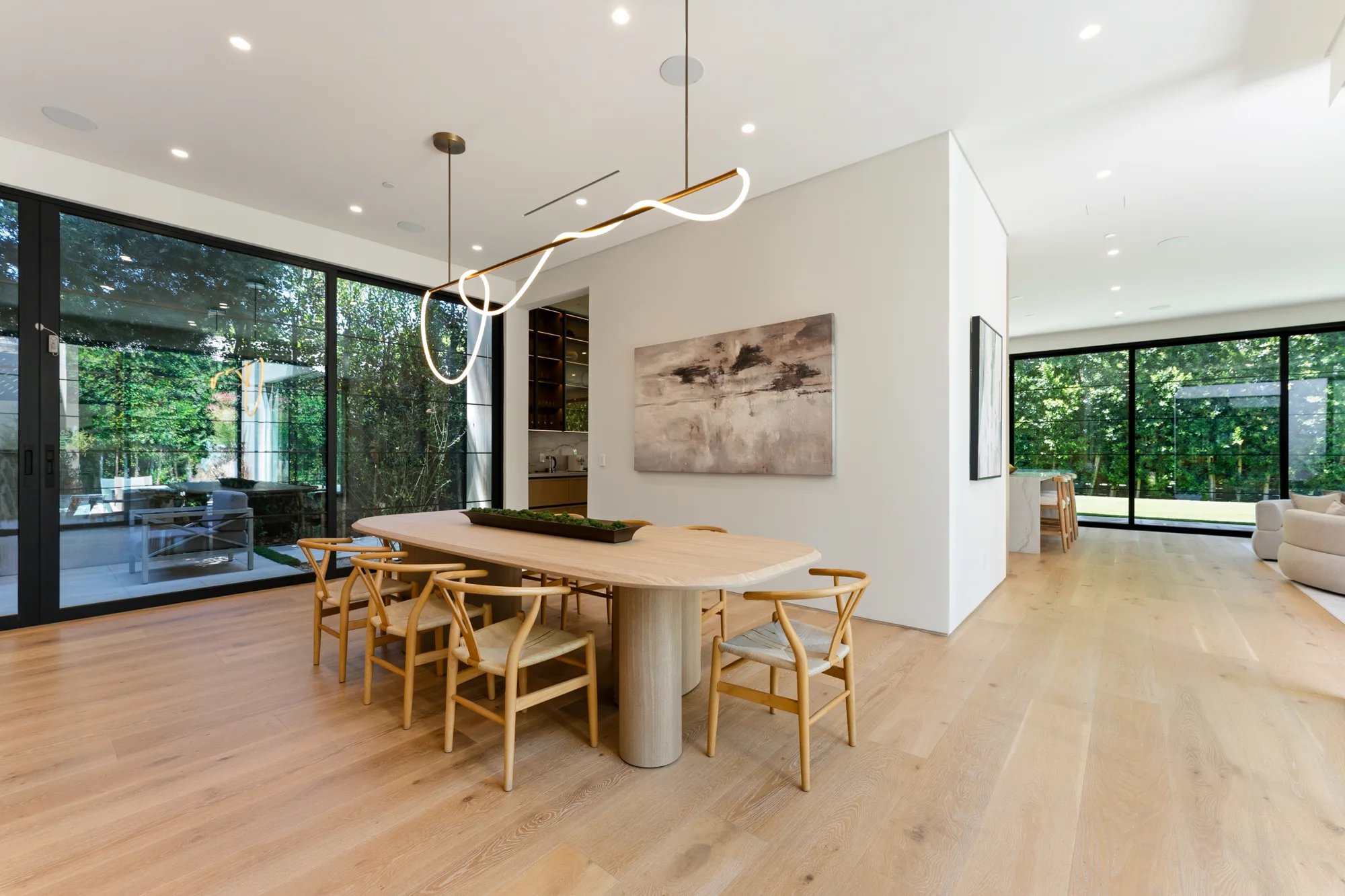 Dining table for eight with wishbone chairs beneath a ribbon light, glass walls on two sides