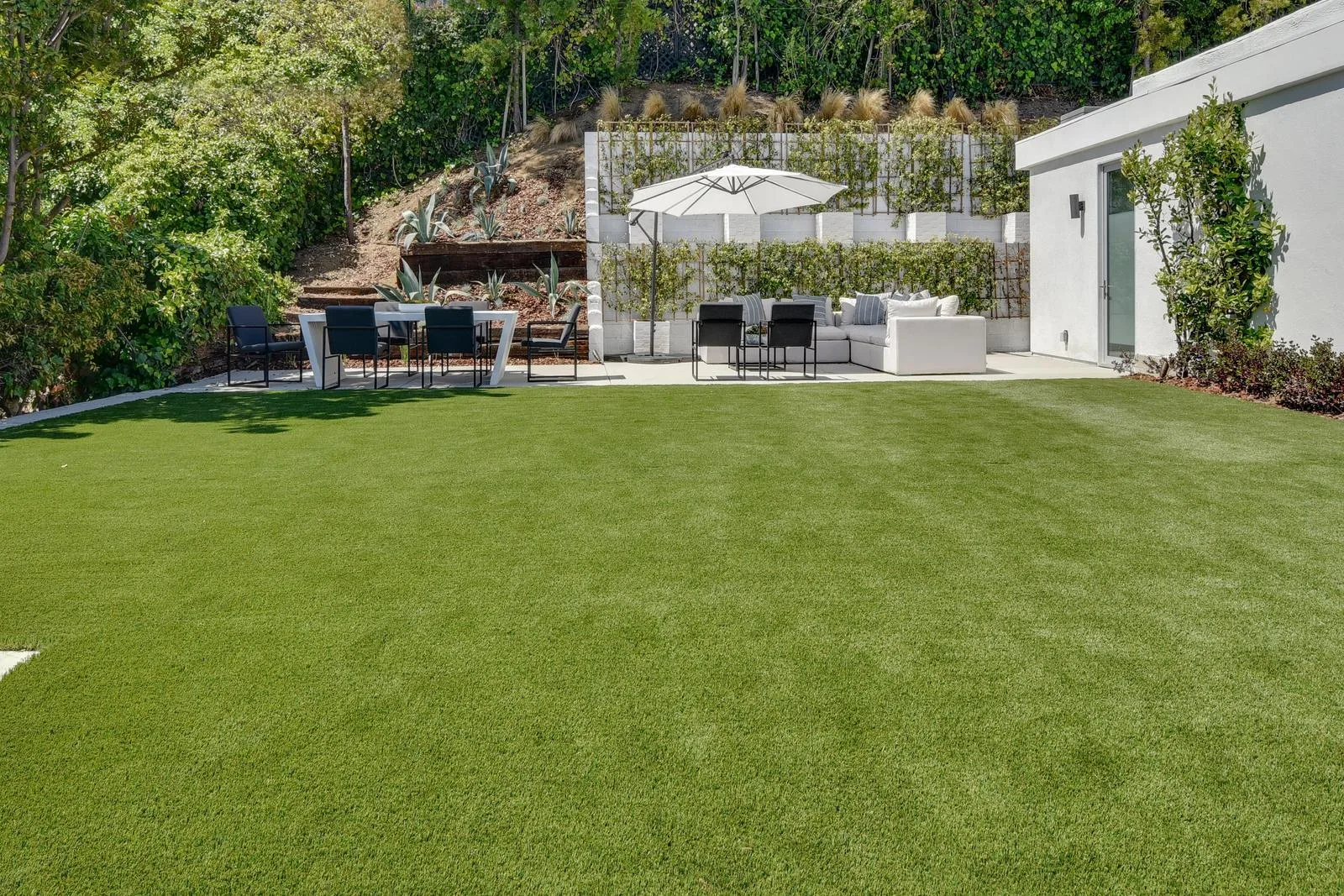Lawn stretching to a shaded dining table and sectional against a planted terraced retaining wall