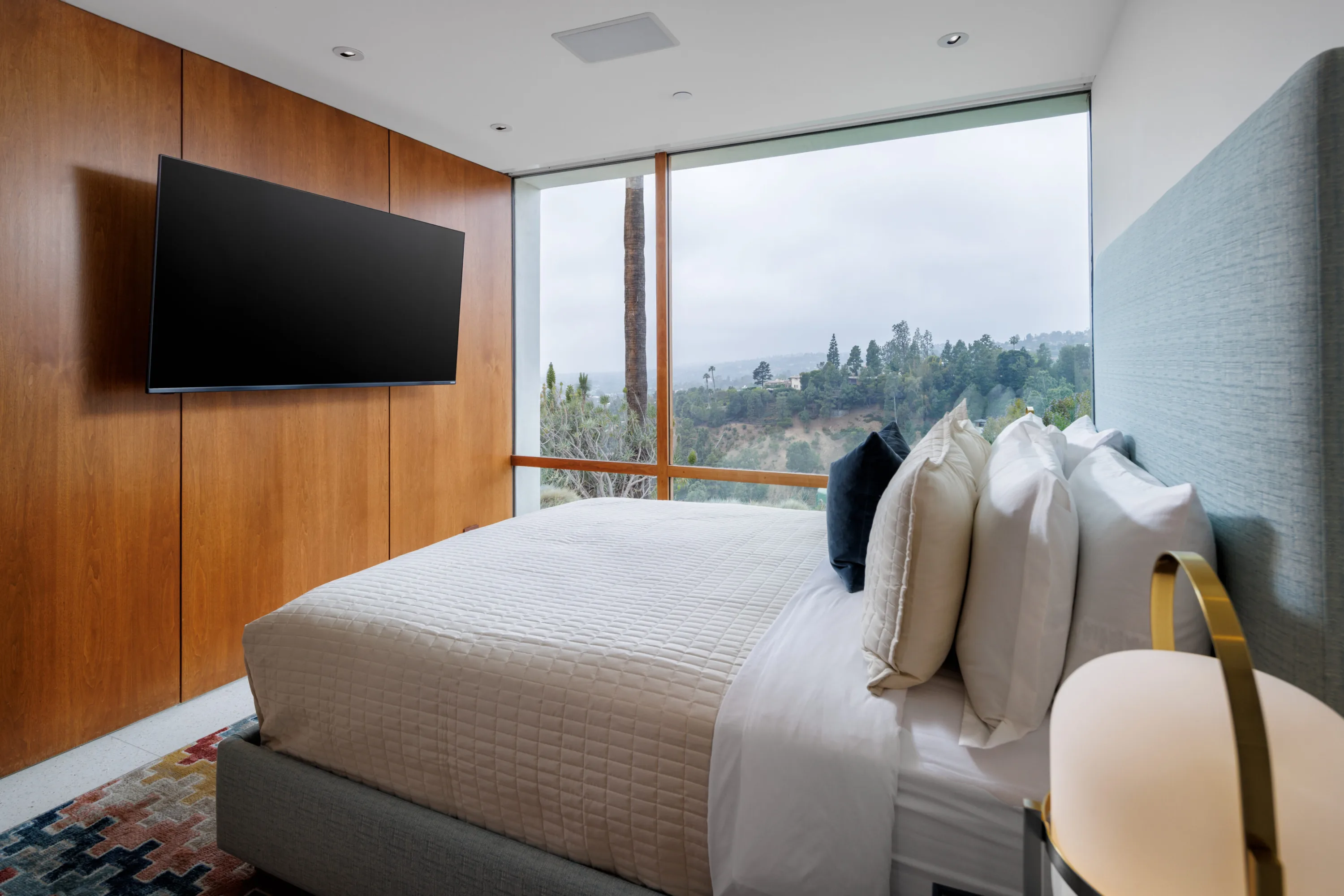 Guest bedroom panelled in warm walnut, floor-to-ceiling glass looking over treetops and ridgeline