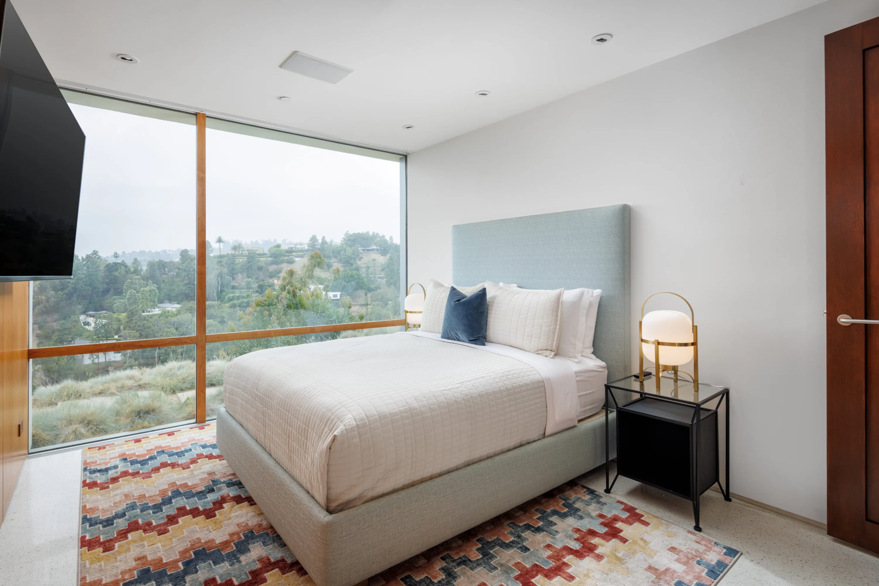Bedroom with pale upholstered headboard, brass globe lamp and window wall over the hillside below