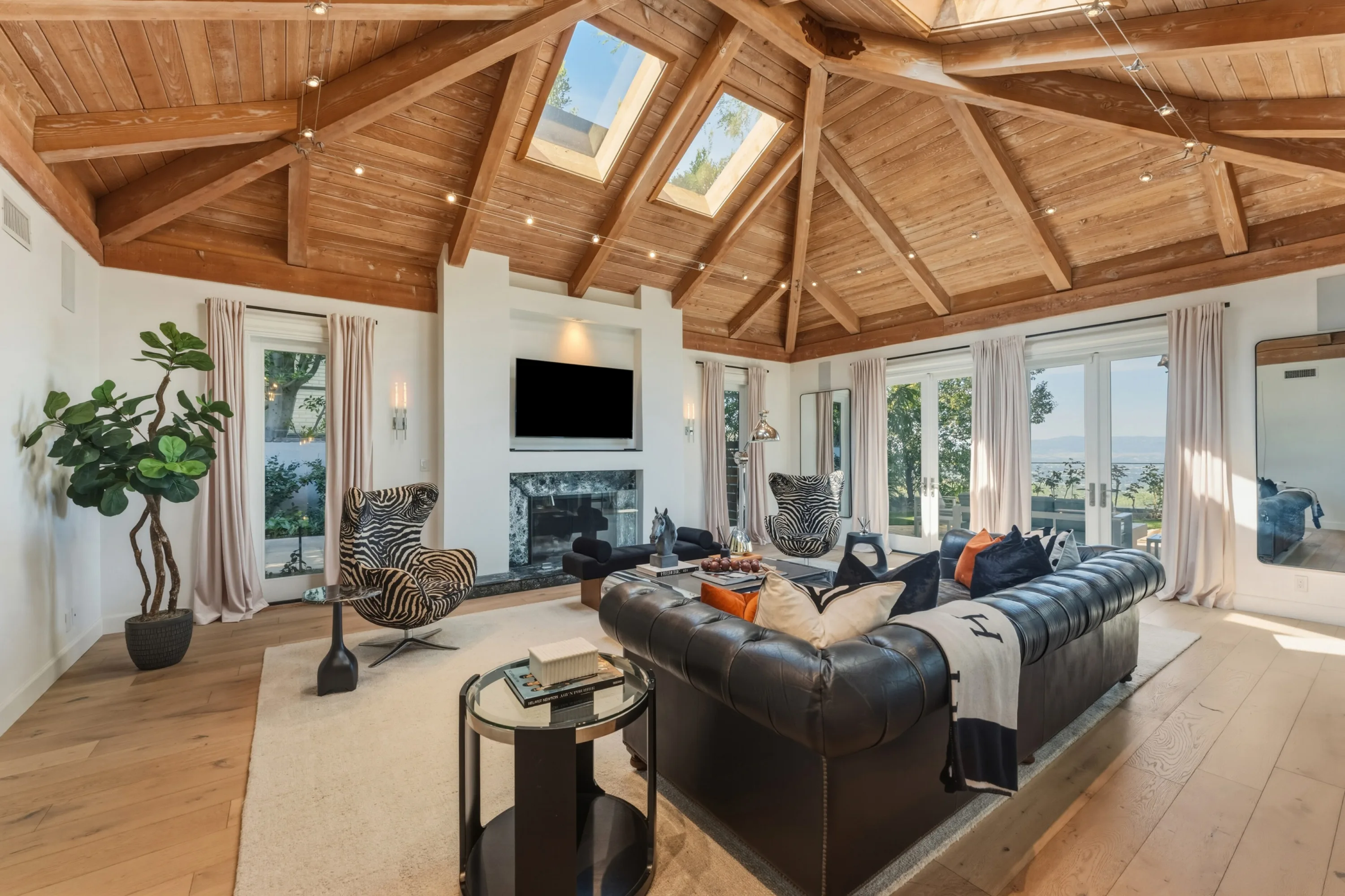 Living room under a vaulted timber-beam ceiling with skylights, stone fireplace and leather Chesterfield