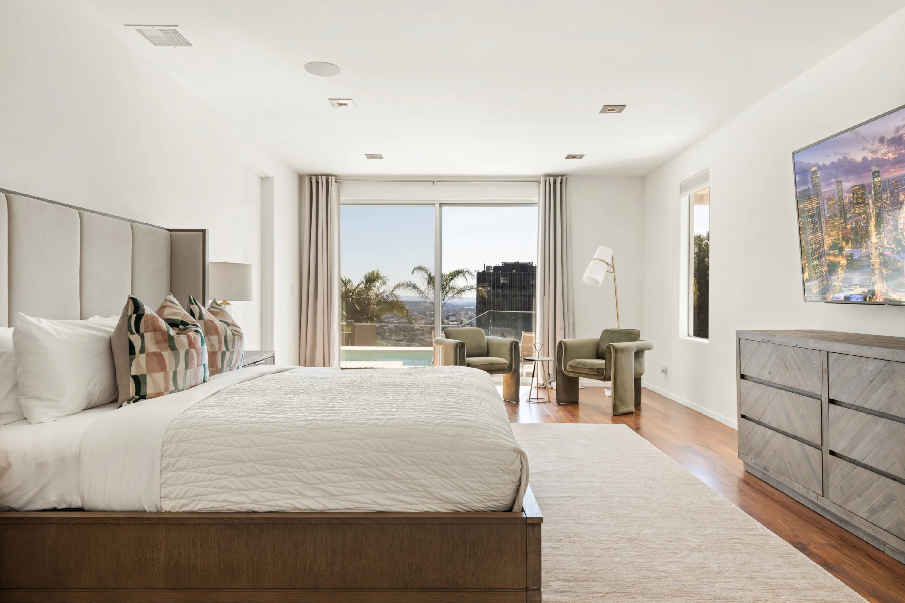 Bedside view across the primary suite to palm-framed balcony and pool water beyond the glass