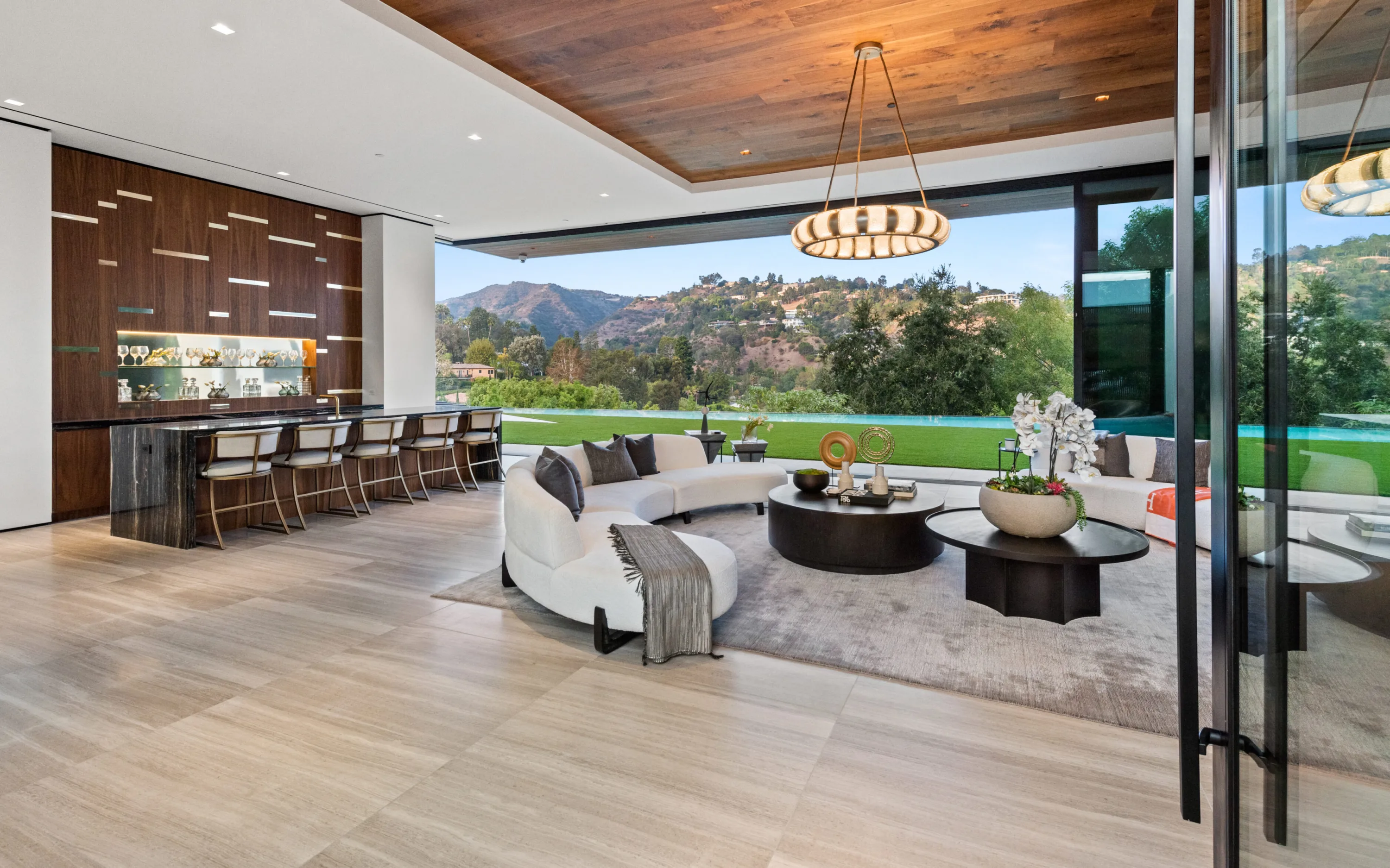 Great room with curved sofas and walnut bar opening fully to lawn, pool and canyon hillside