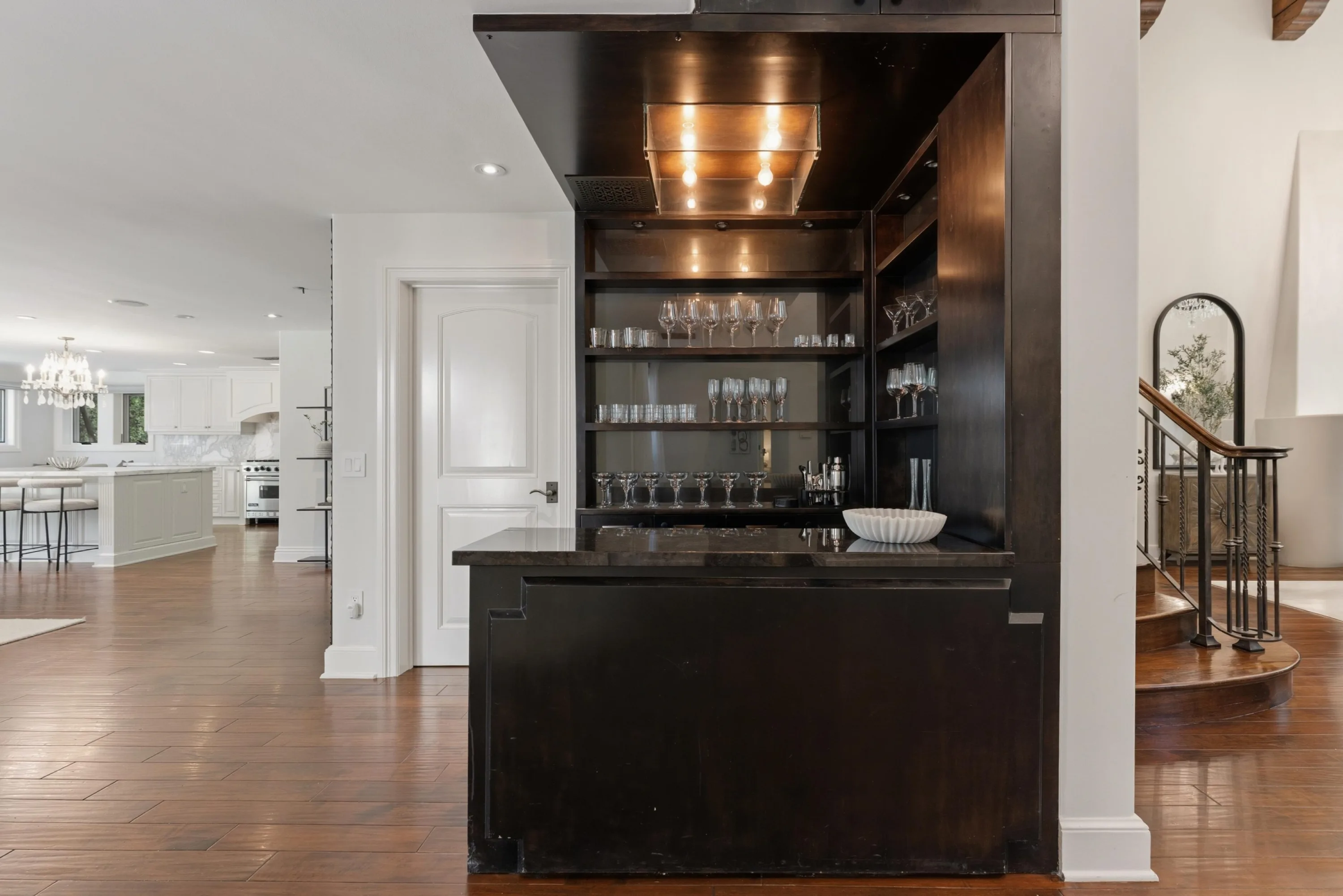Dark-wood wet bar with lit glass shelving and stemware, open to the kitchen and staircase
