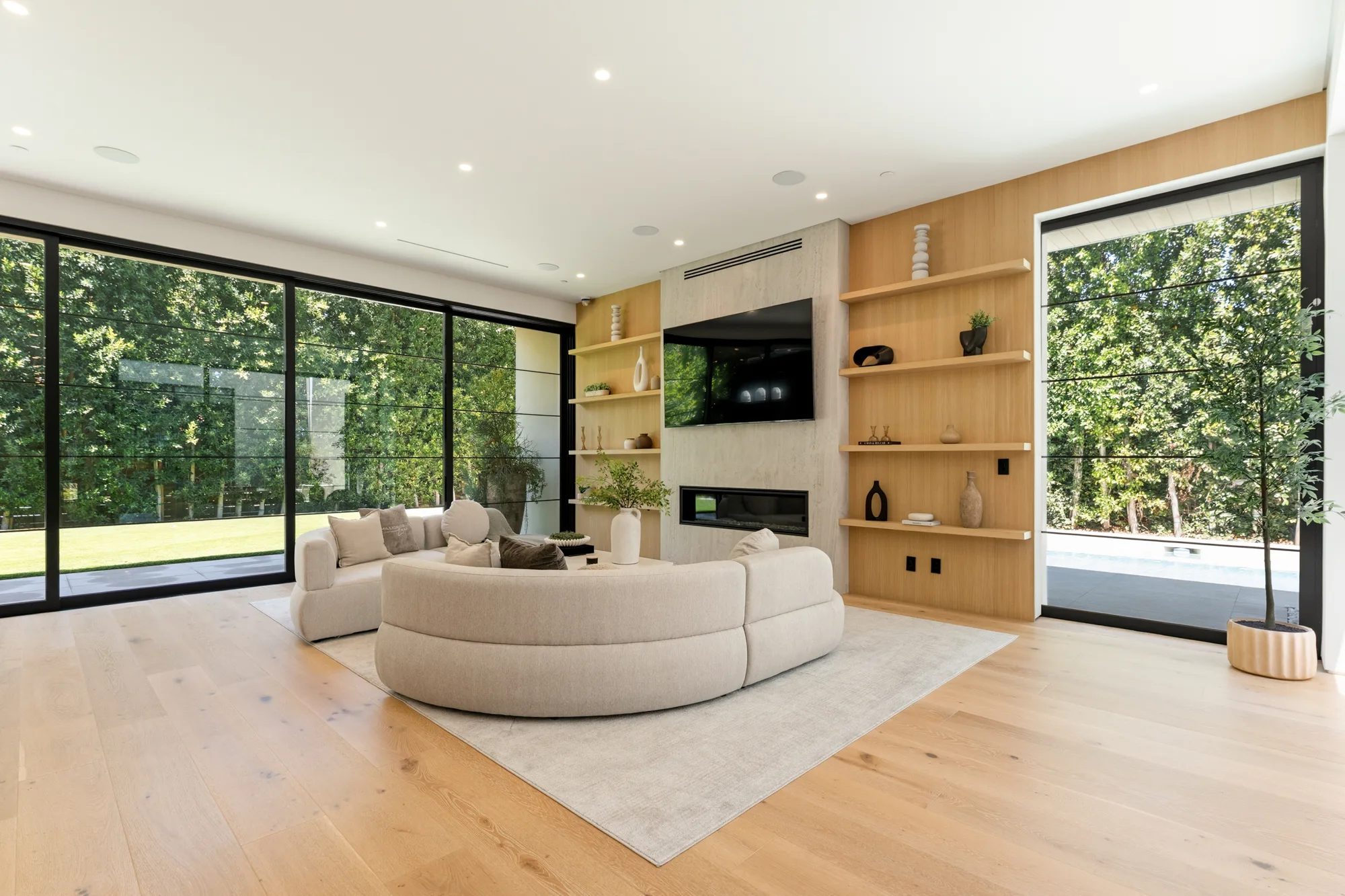 Family room with curved sectional facing a fireplace, TV and oak shelving, lawn visible through glass