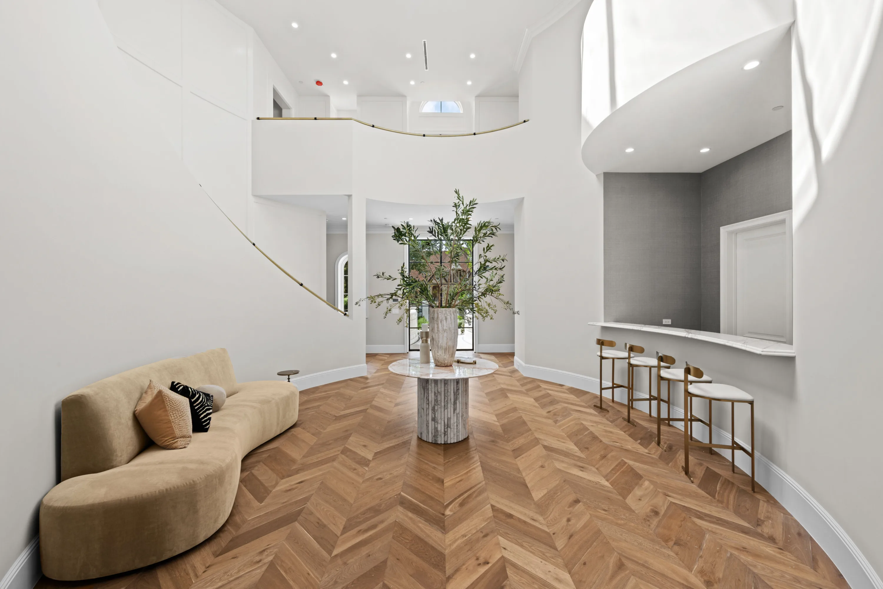 Double-height entry hall with sweeping stair, chevron floors, centre table and brass bar stools at a stone counter