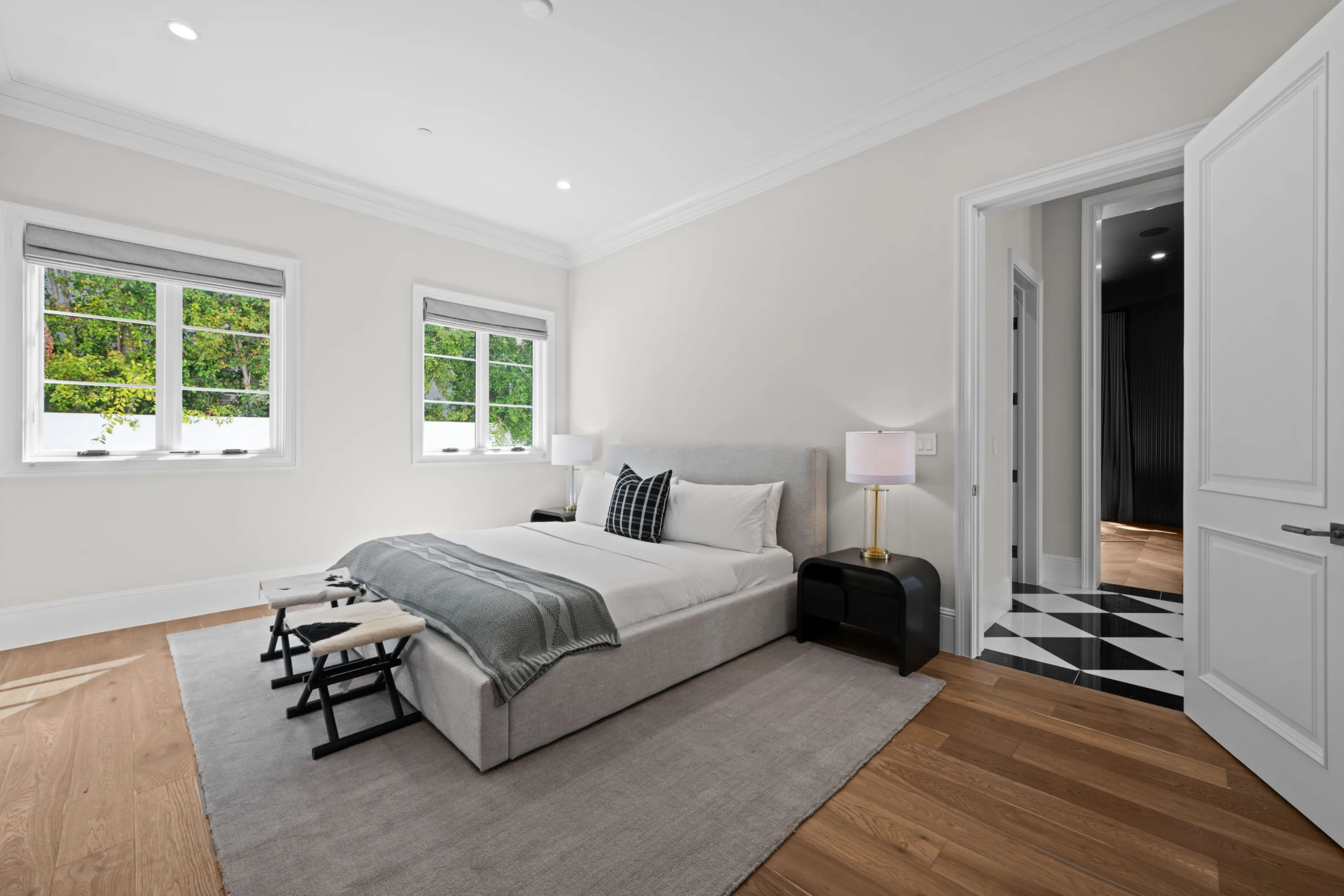 Bright bedroom with upholstered bed, twin windows over greenery and a doorway to a checkerboard-floored bath