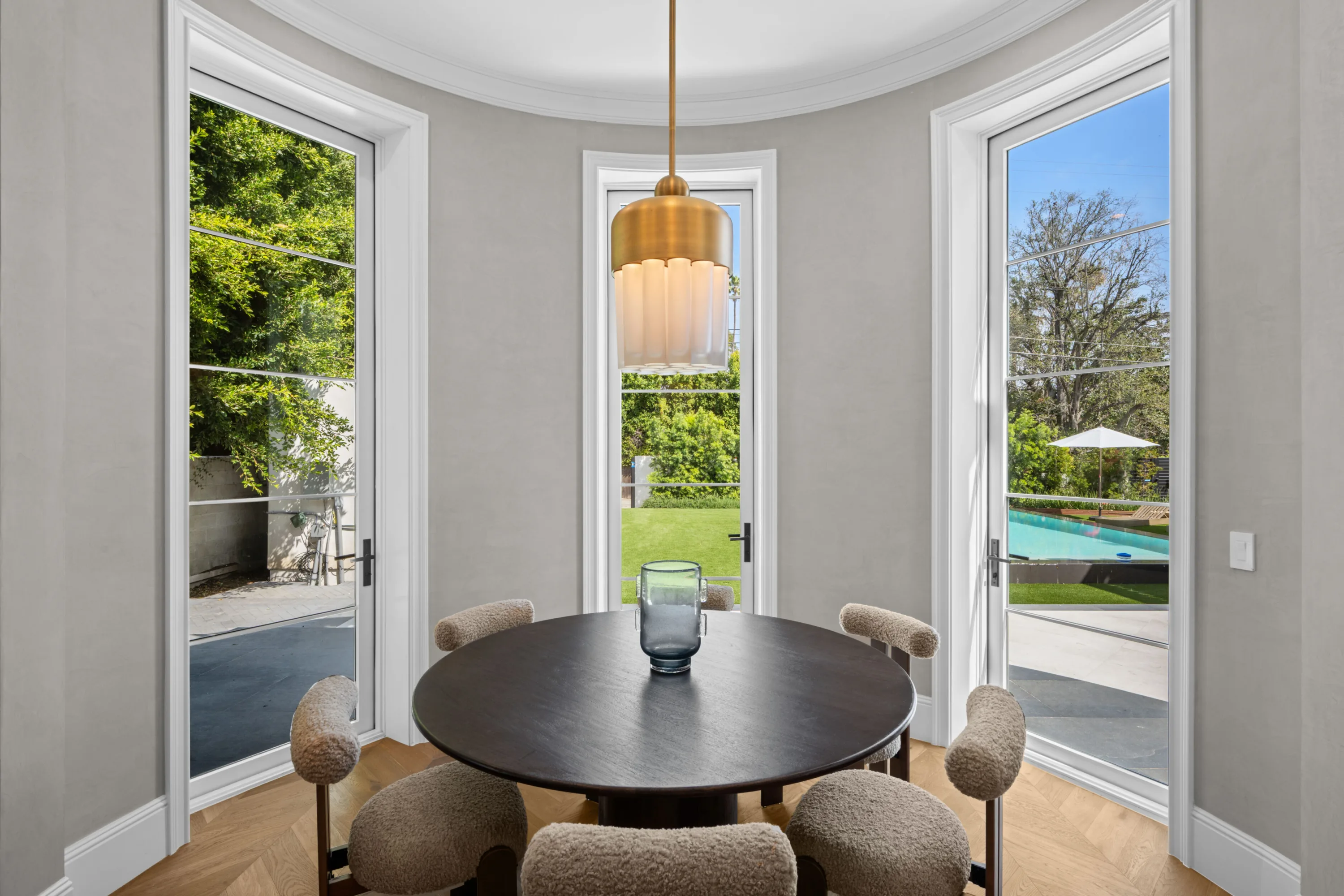 Curved breakfast nook with round table, bouclé chairs and French doors framing the lawn and pool