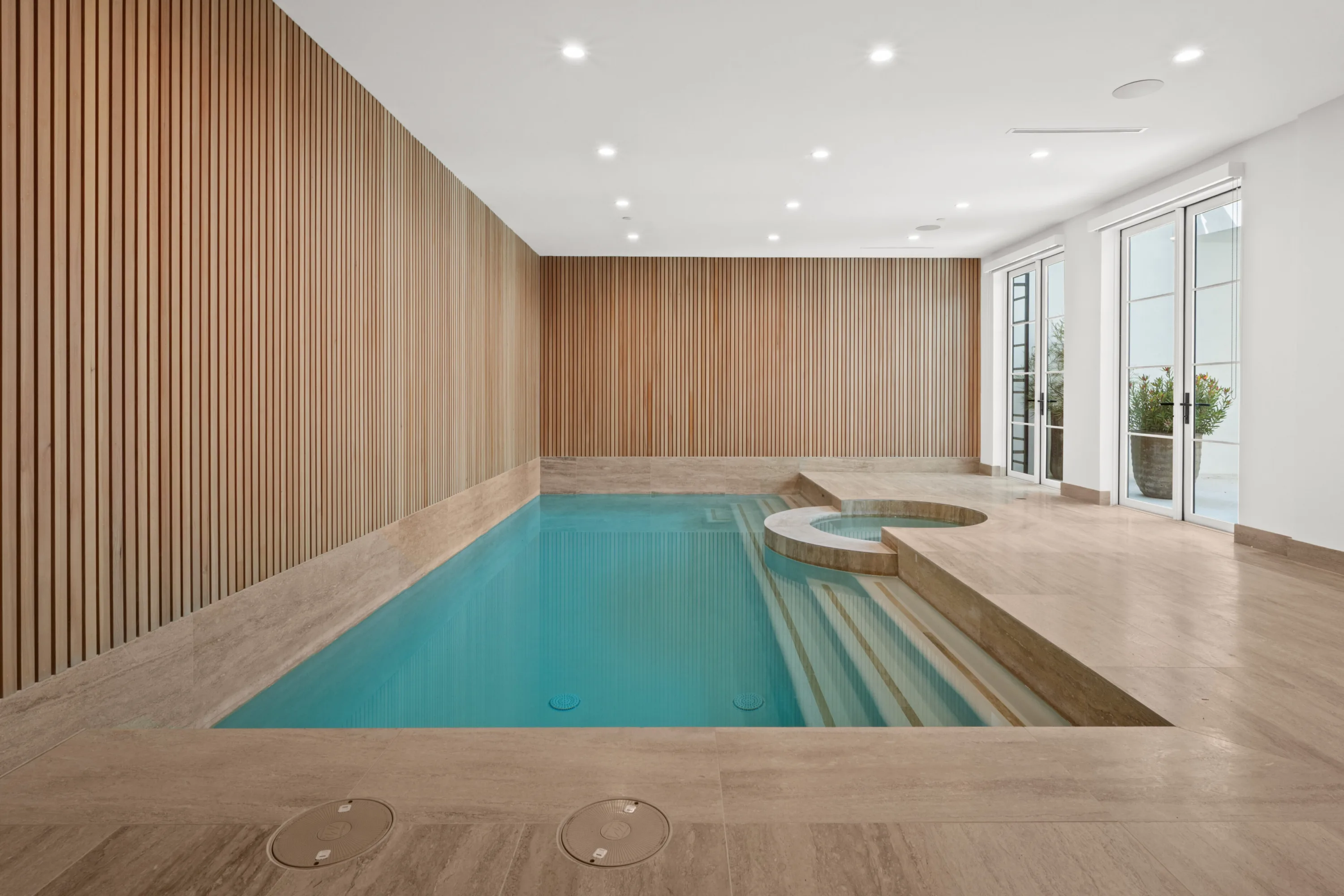Indoor pool and inset spa lined in travertine, with slatted timber walls and doors to a planted courtyard