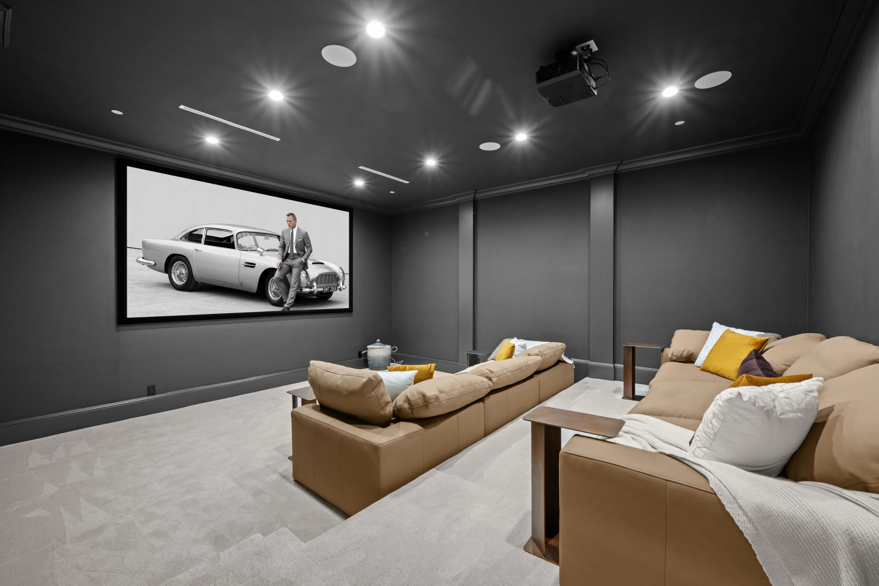Home cinema in charcoal with tan leather lounges facing a projection screen showing a man beside a vintage sports car