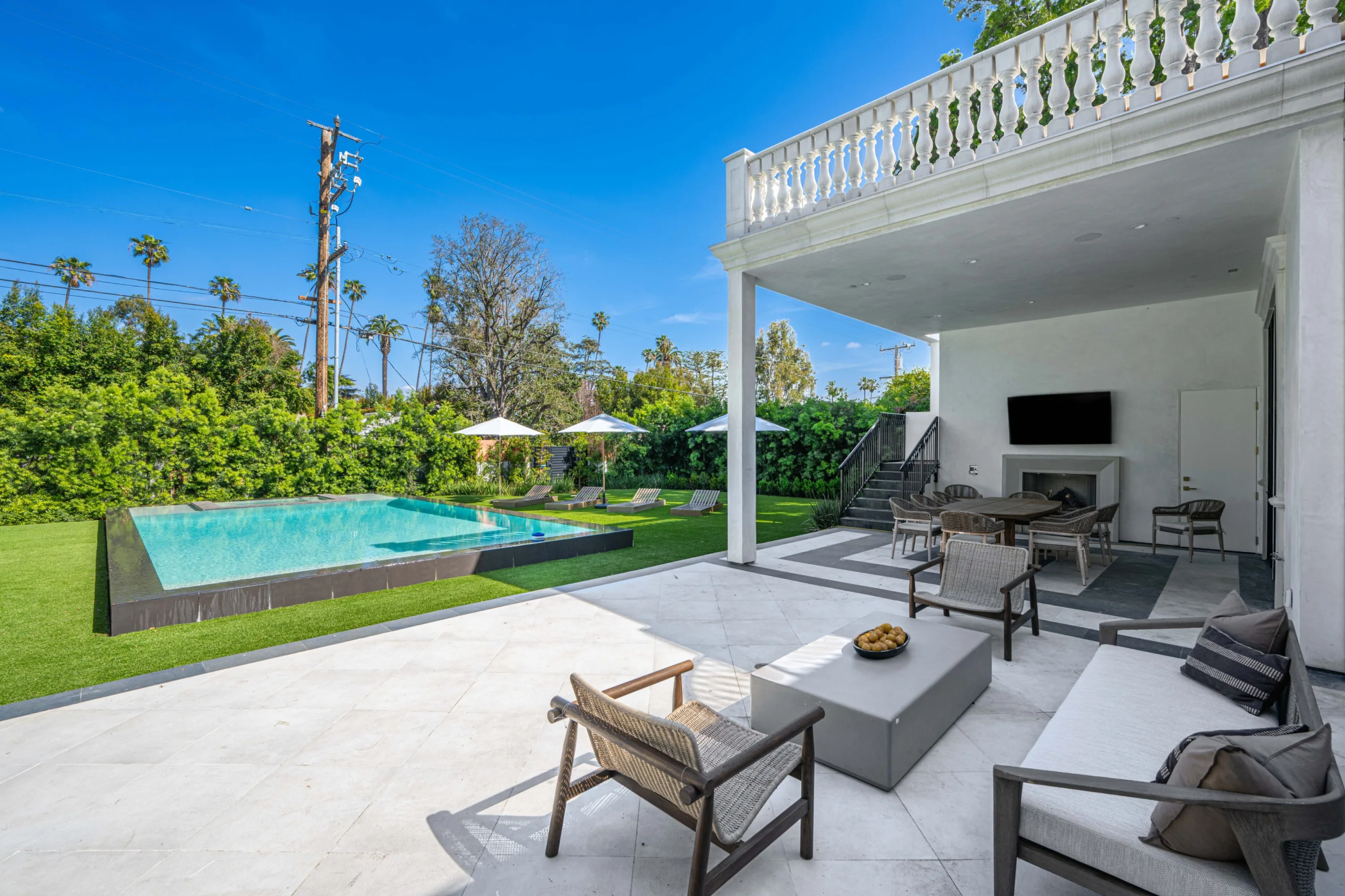 Covered loggia with fireplace and dining set looking across travertine terrace to the pool and lawn