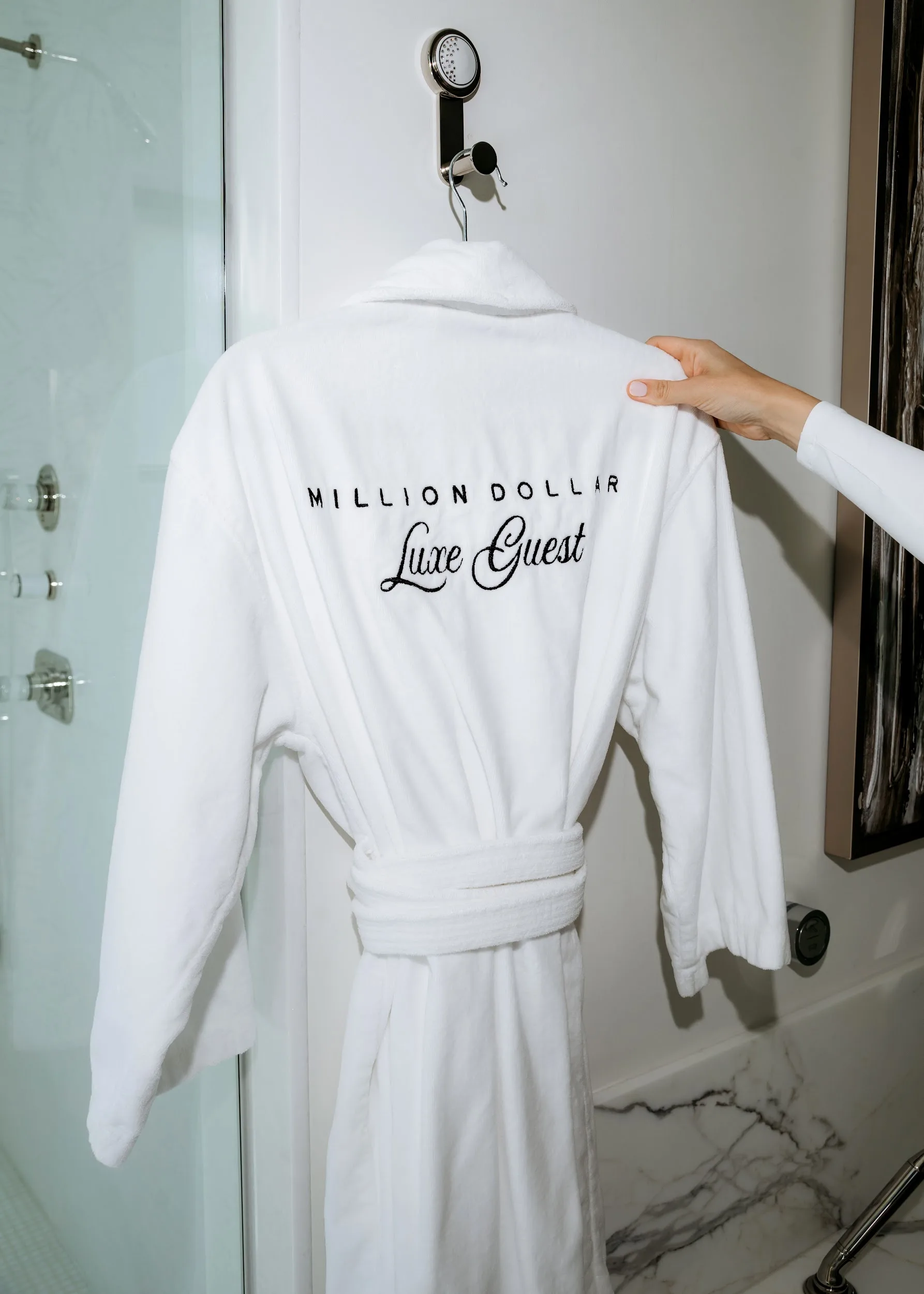 A hand lifting a white robe embroidered "Million Dollar Luxe Guest" on a hook in a marble guest bathroom