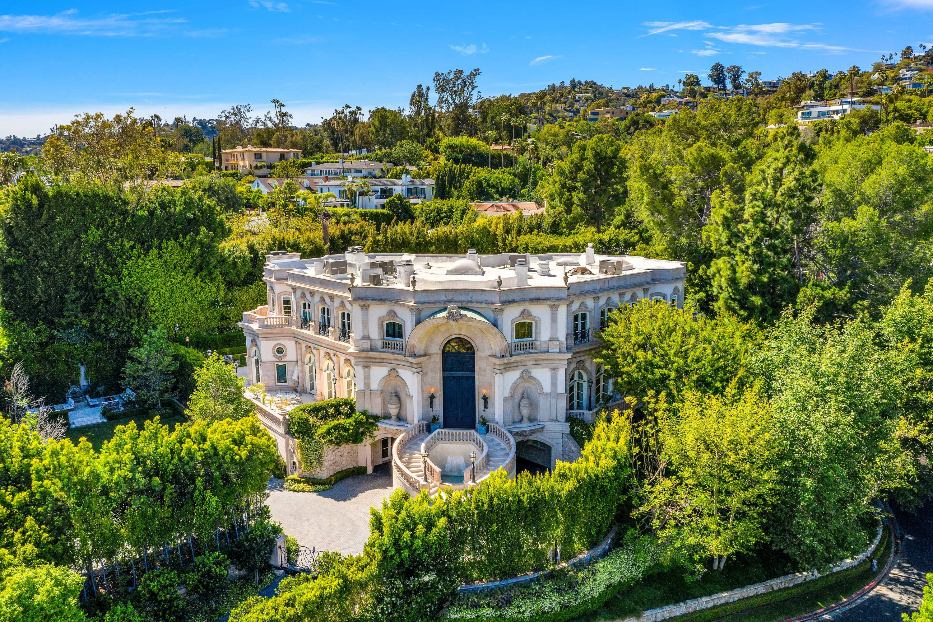 Aerial view of a grand curved white mansion with dual sweeping staircases to a circular fountain court, set among dense hillside greenery