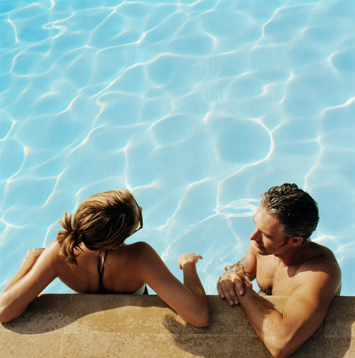 Two guests resting against the stone edge of a sunlit swimming pool, sunglasses pushed into wet hair
