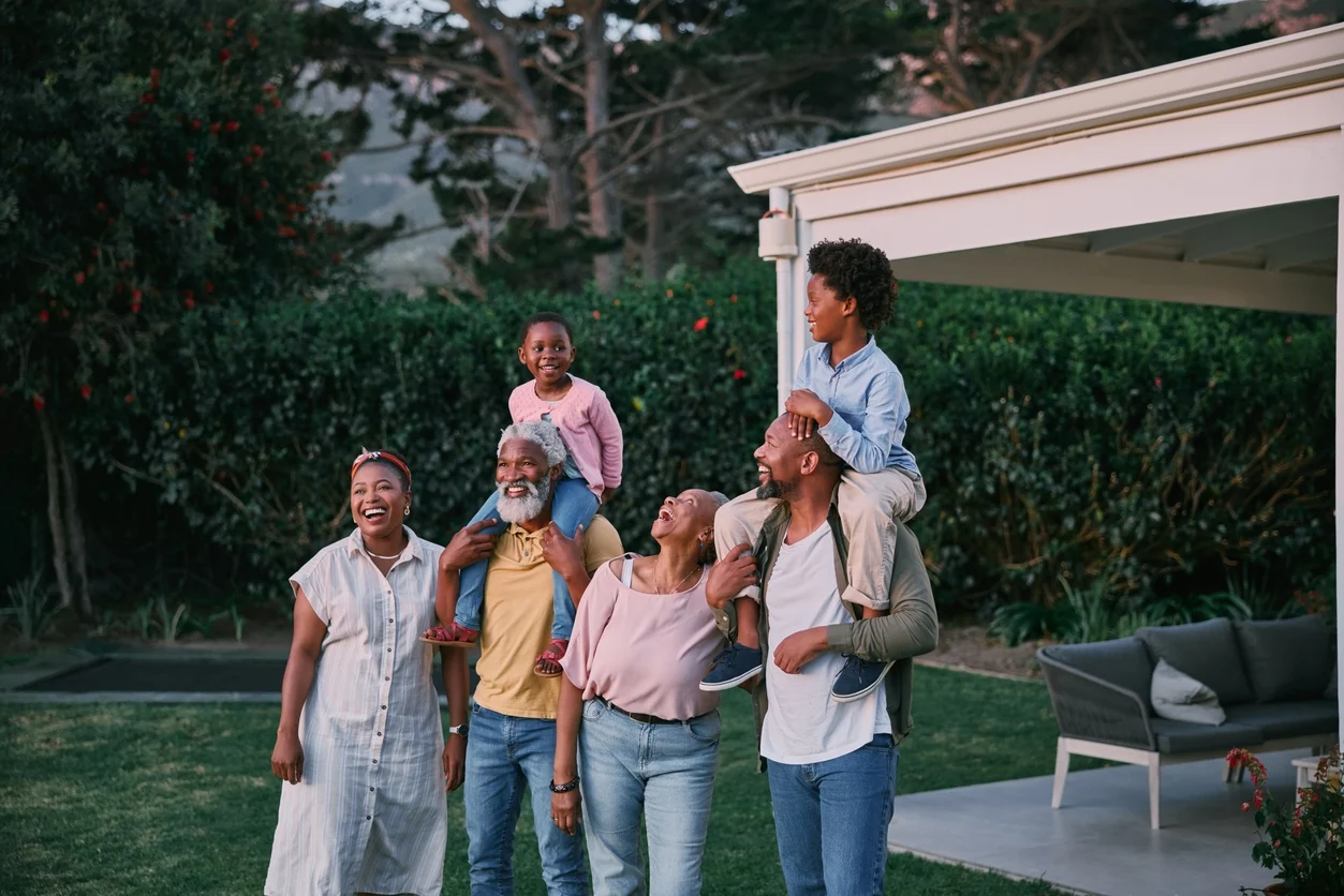 Six guests of three generations laughing on the lawn, two children on shoulders, beside a covered patio with sofa