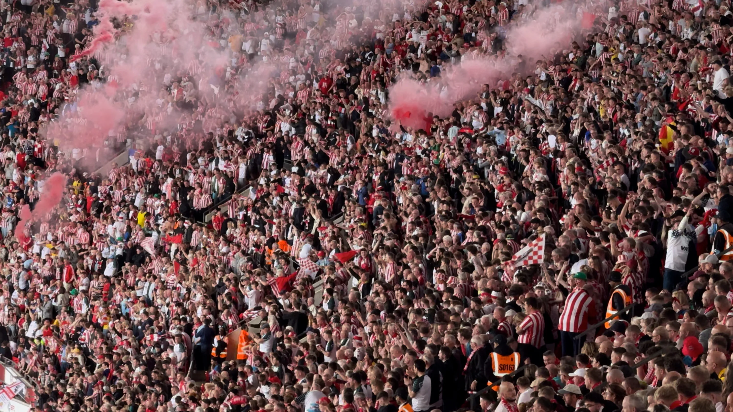 Packed stadium stand of fans in red-and-white stripes waving flags amid drifting pink flare smoke