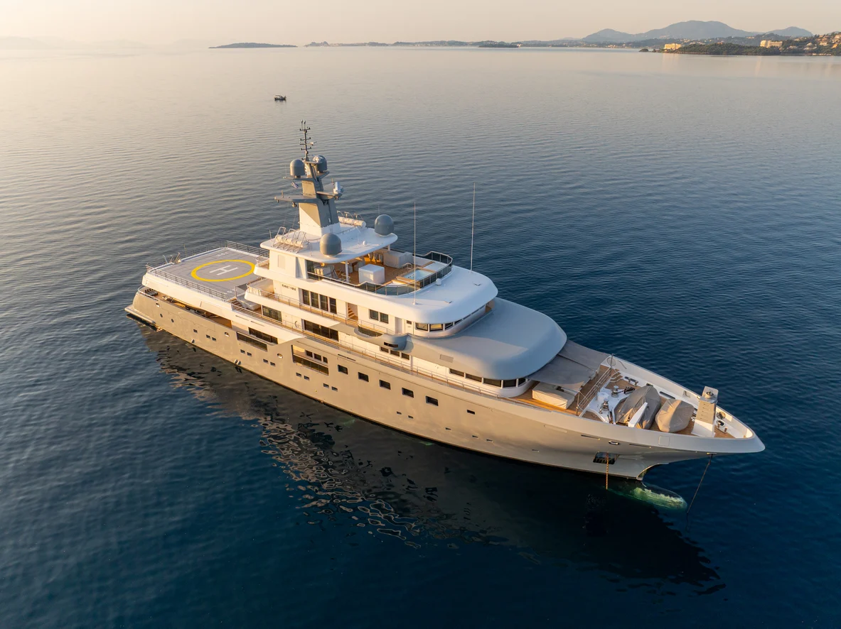 Aerial view of a superyacht anchored in calm water at golden hour, helicopter pad on the bow and coastal hills in the distance