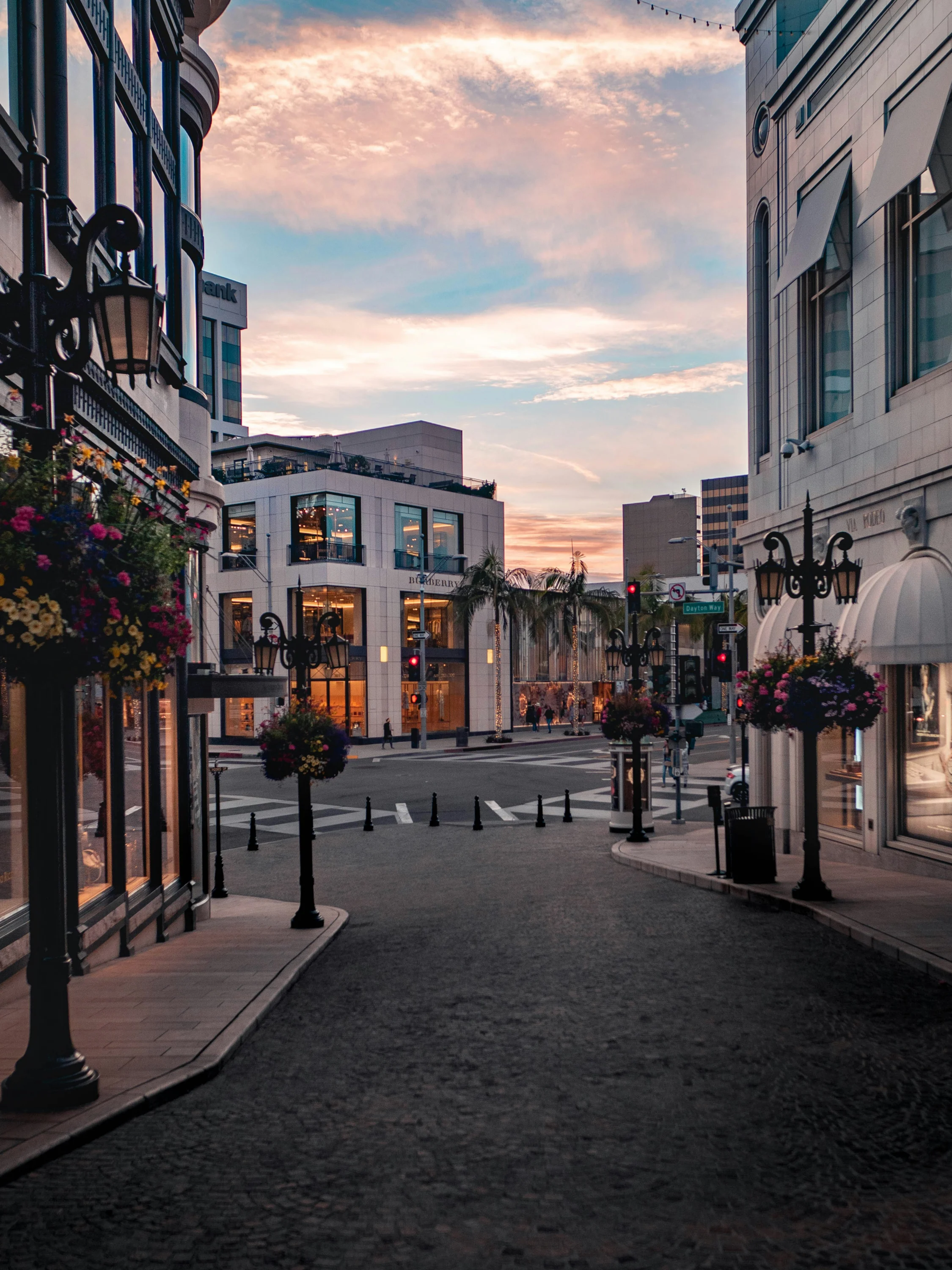 Dusk view down an empty Rodeo Drive, flower-draped lamp posts and boutique storefronts under a pink and blue sky