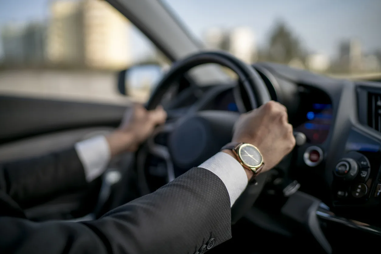 Driver in a dark suit and gold watch, both hands on the wheel, city buildings blurred through the windshield